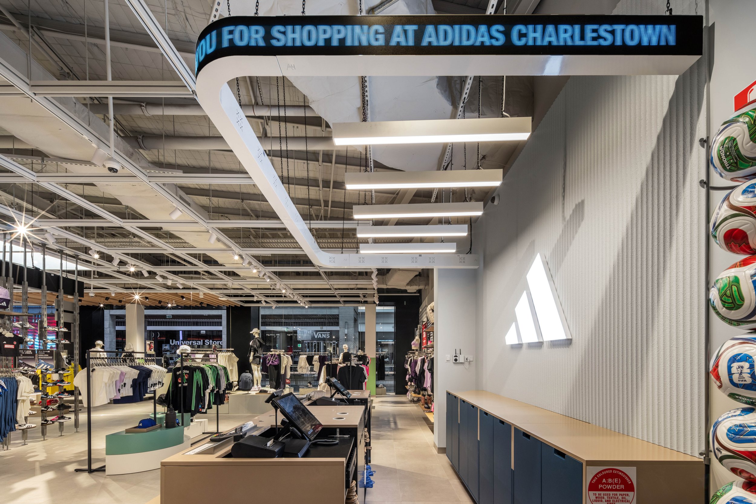 Inside a retail store with clothing displays, checkout counters, and spherical shelves with sports balls on the right. An LED sign on the ceiling reads 'THANK YOU FOR SHOPPING AT ADIDAS CHARLESTOWN.'