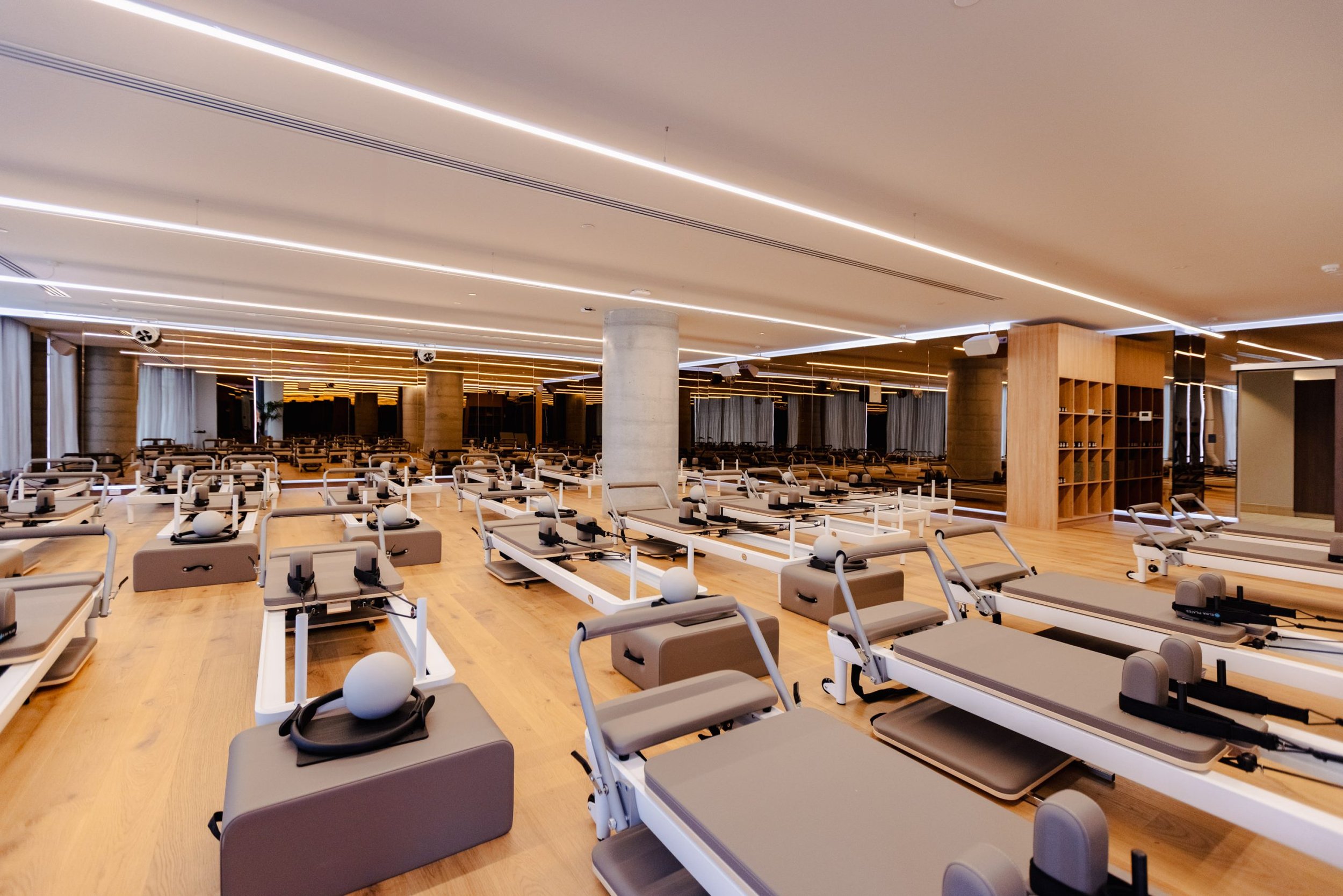 Gym studio with multiple Pilates reformer machines arranged in rows, large windows with curtains, modern lighting, wooden floors, and wooden shelving.