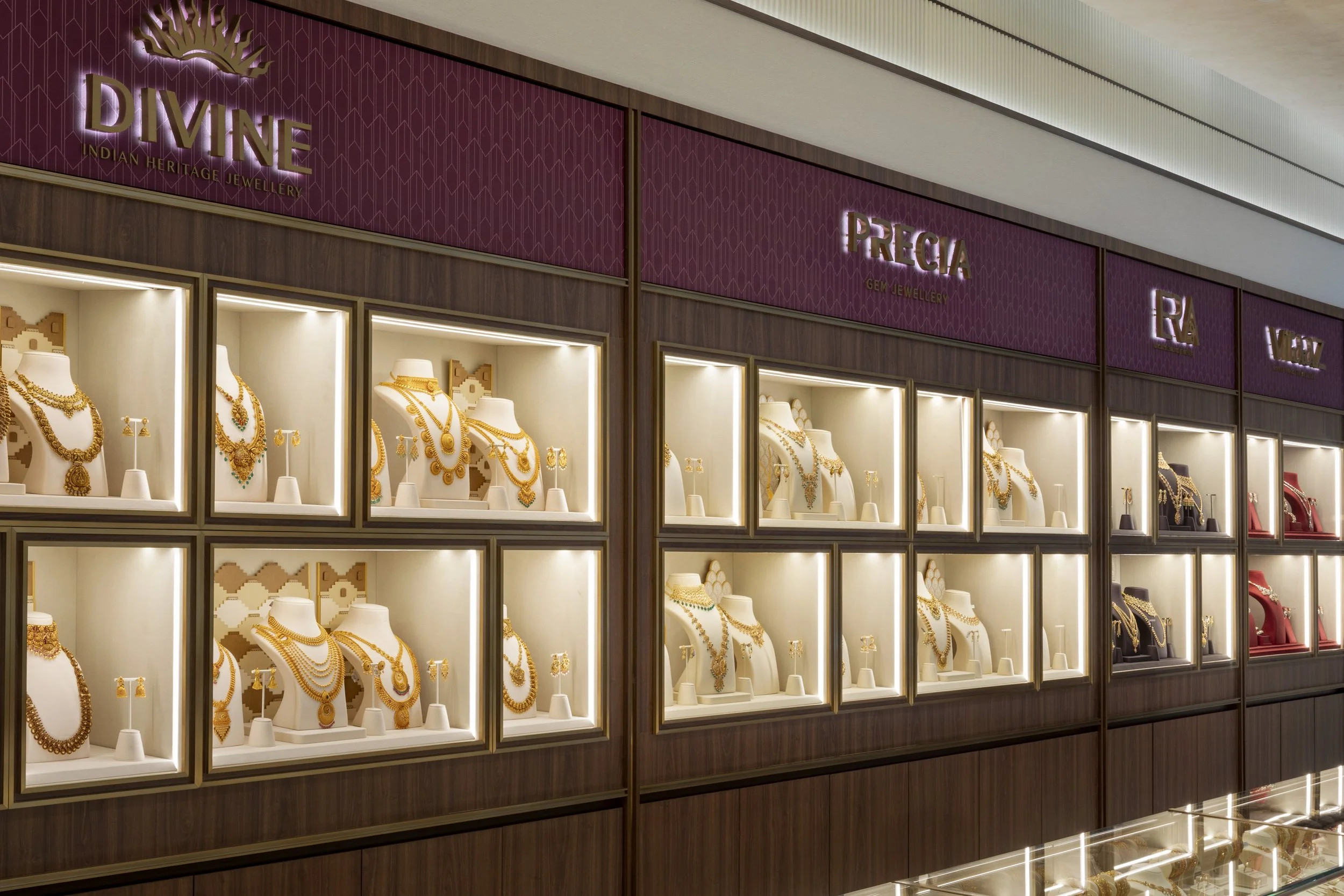 Display case filled with gold jewelry, including necklaces, earrings, and pendants, in a jewelry store. The store has signs with the brands 'DIVINE' and 'PRECIA' on a purple background.