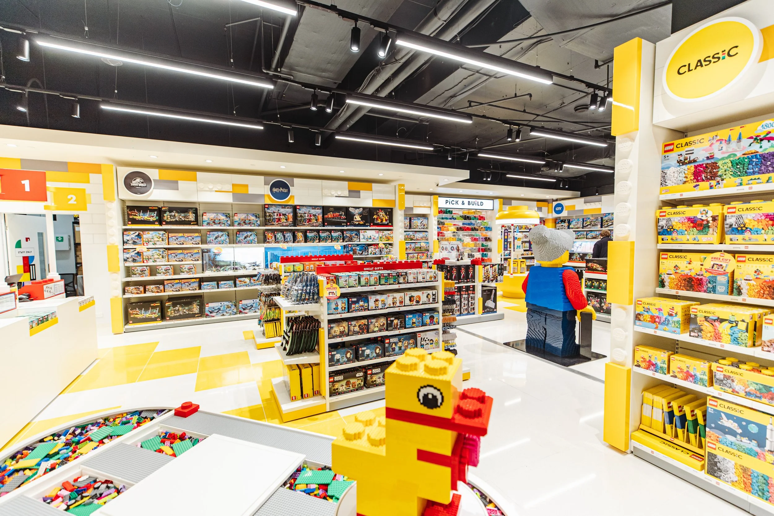 Lego store interior displaying shelves with Lego sets and building blocks, including notable sets like Lego Harry Potter and Lego Classic, with a central Lego statue and bright, modern lighting.