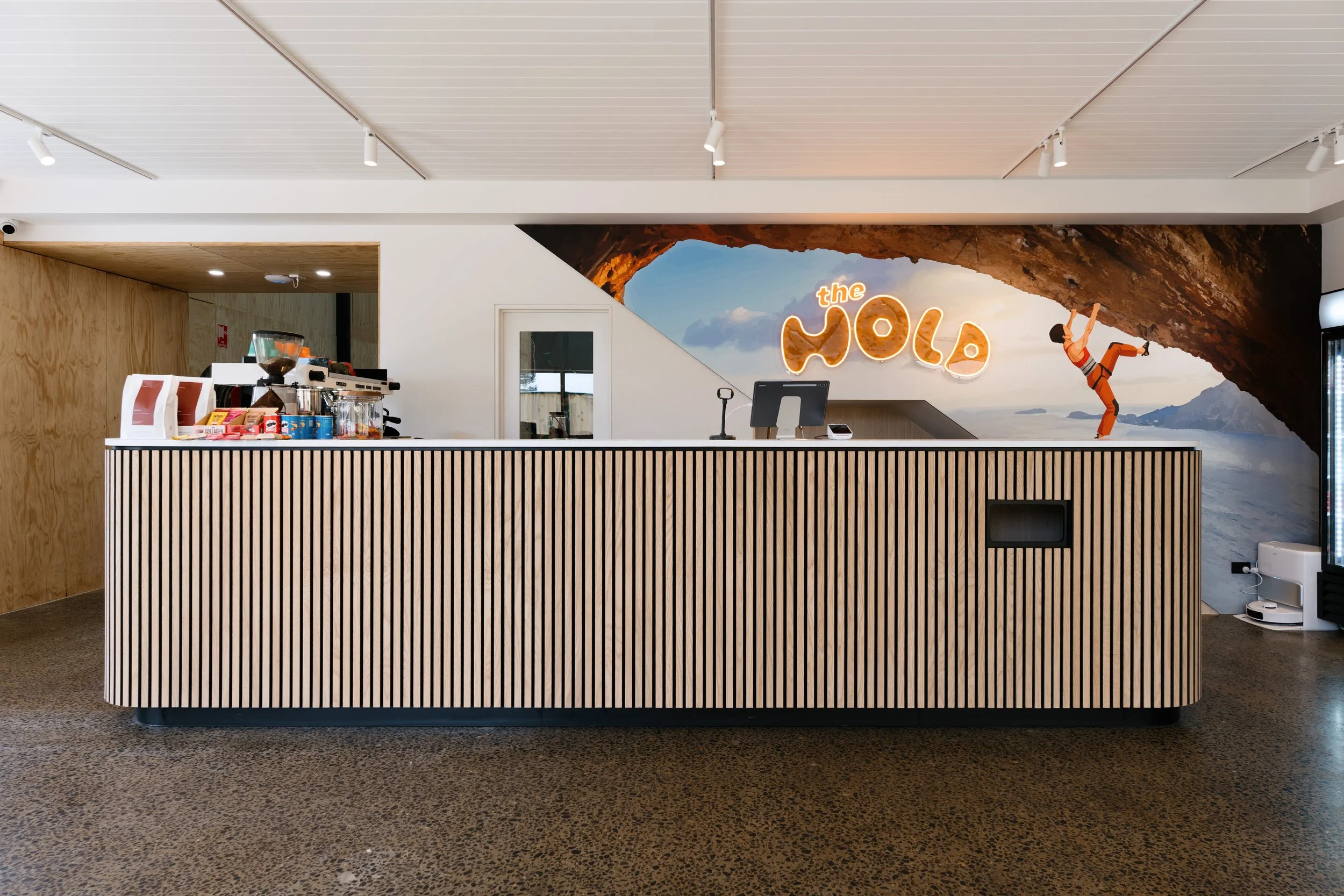 A reception desk with a curved wooden front panel in an indoor space, featuring a mural of a rock formation with a climber, clouds, and mountains in the background, and a neon sign that says 'the Wola'.