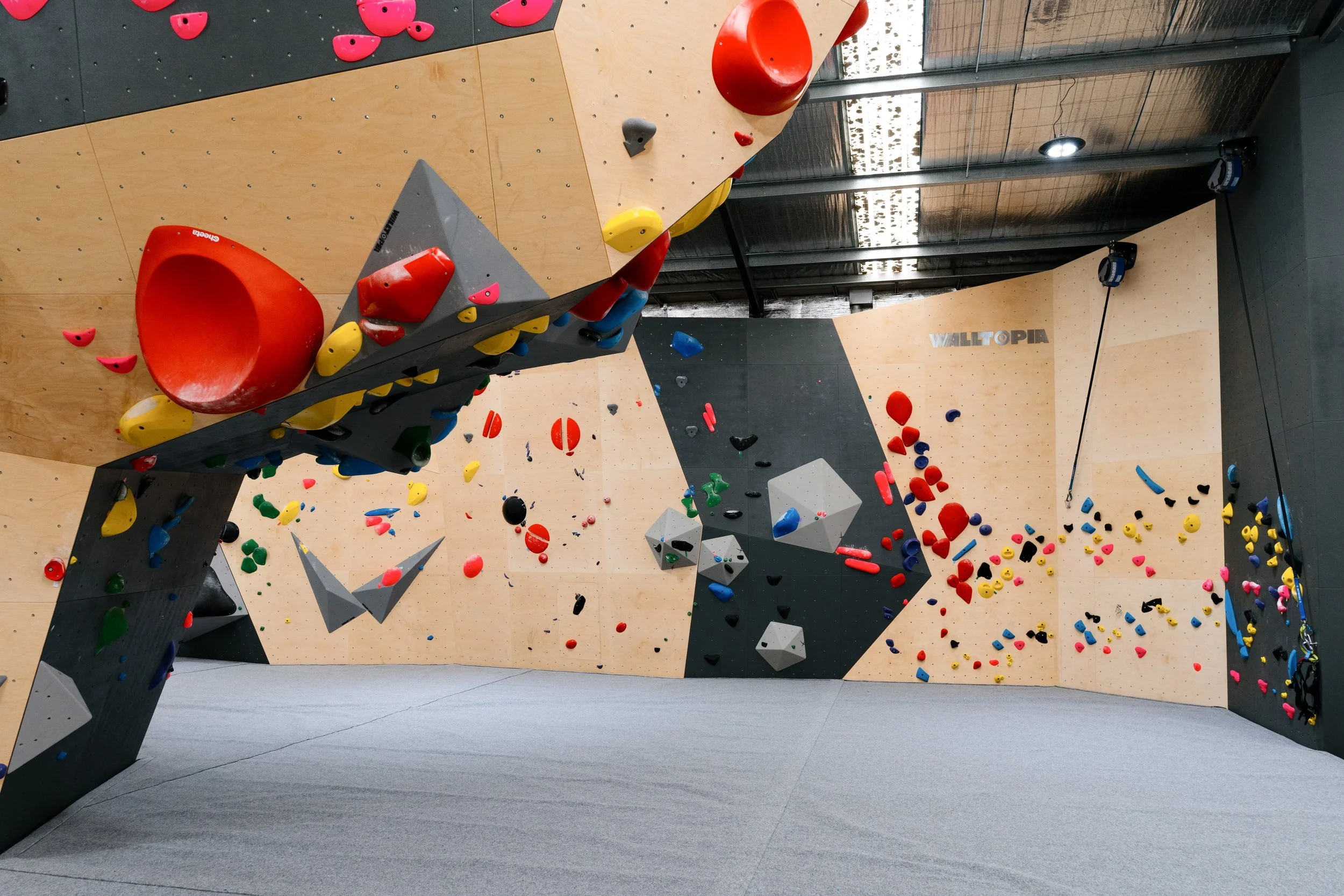 Indoor rock climbing gym with various colorful holds, including large red and yellow shapes and smaller black, green, blue, and pink holds, on different climbing walls.