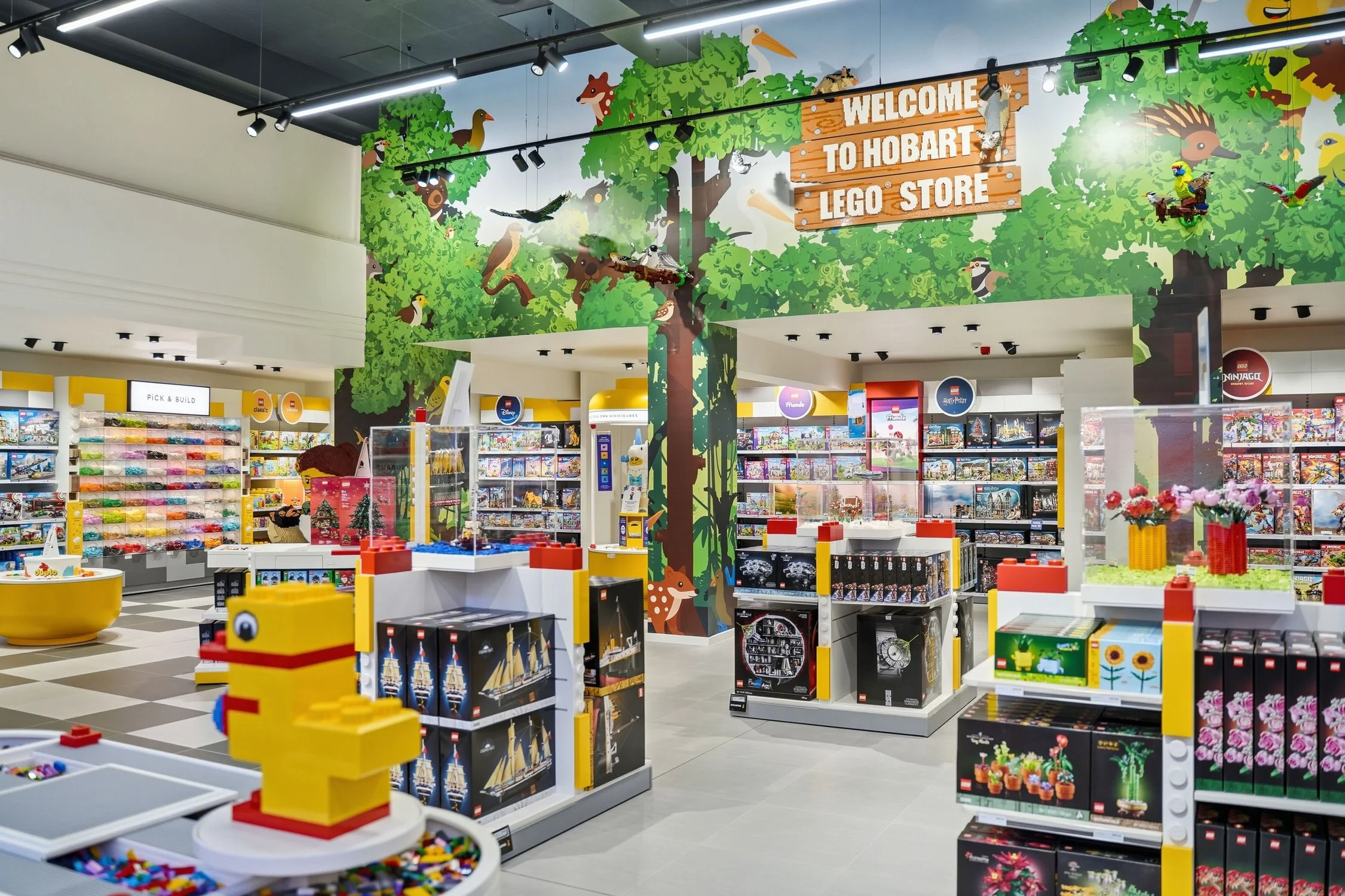 Inside a LEGO store with shelves of LEGO sets, colorful display tables, and a large wall mural featuring a forest scene with animals and a sign.