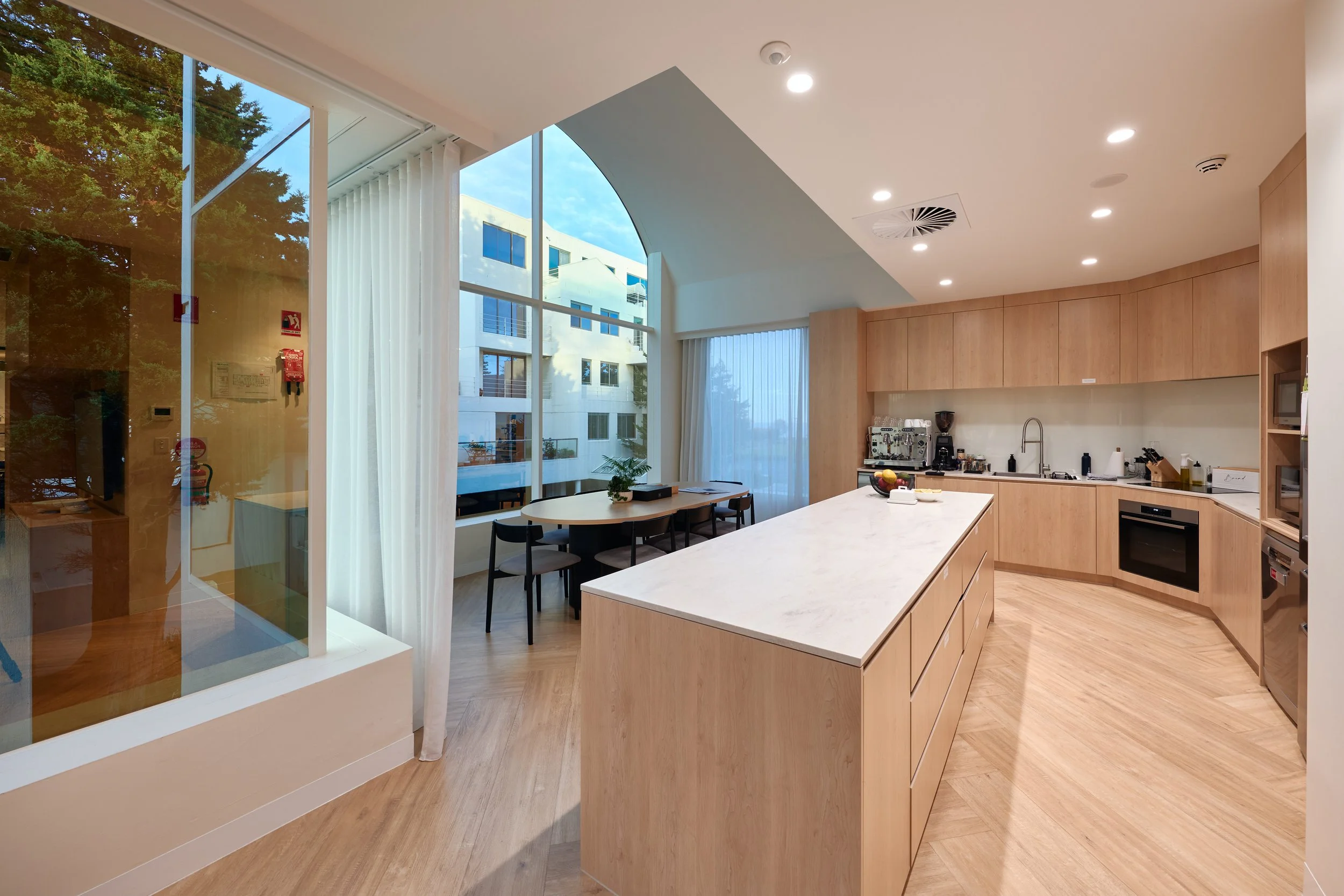 Modern kitchen with wood cabinets, white marble island, and large windows letting in natural light, overlooking a courtyard with a tree.