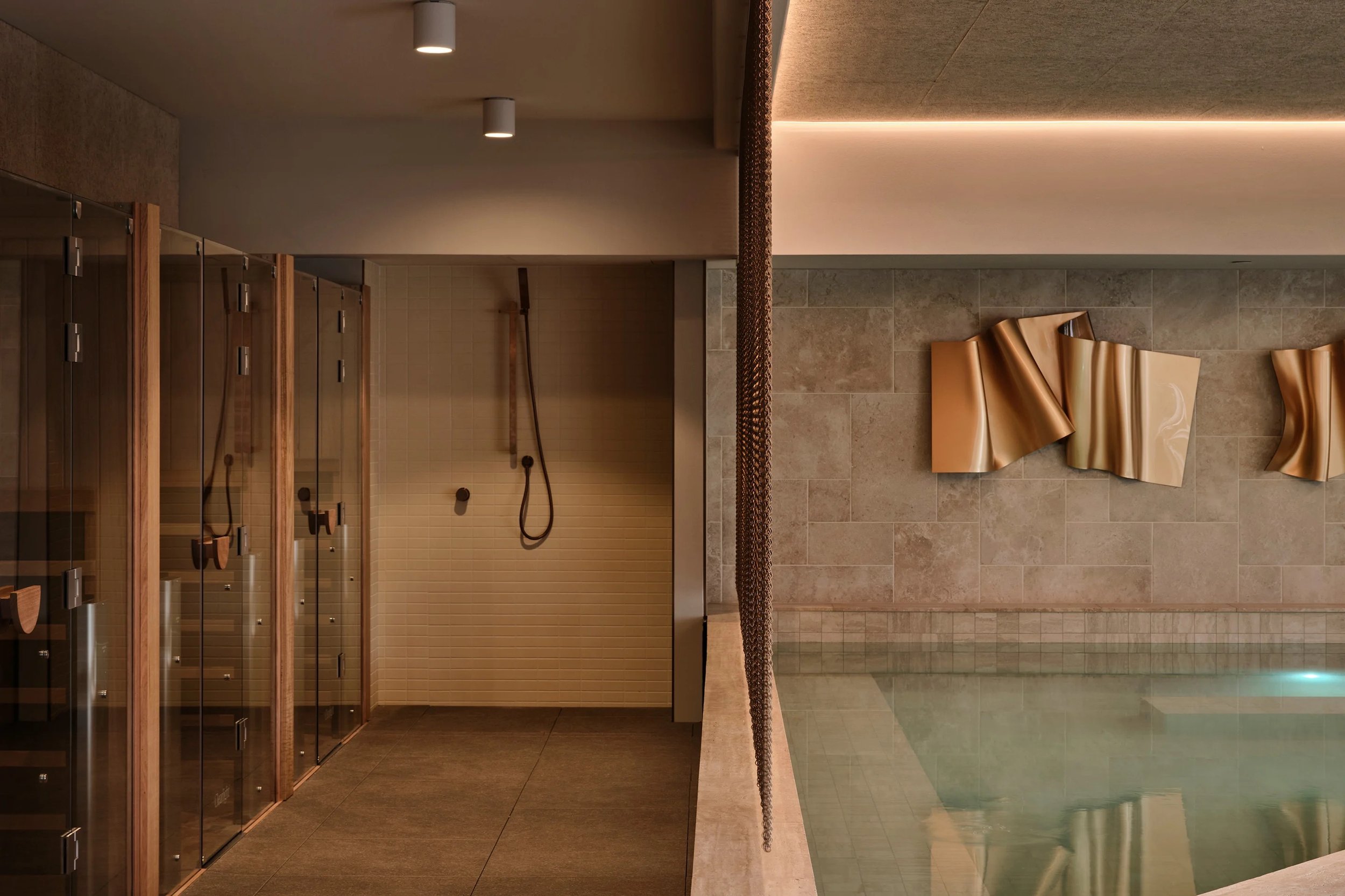 A modern indoor wellness or spa facility with a sauna area, wooden lockers, a shower area with handheld showerheads, and abstract metal wall art fixtures on tiled walls.