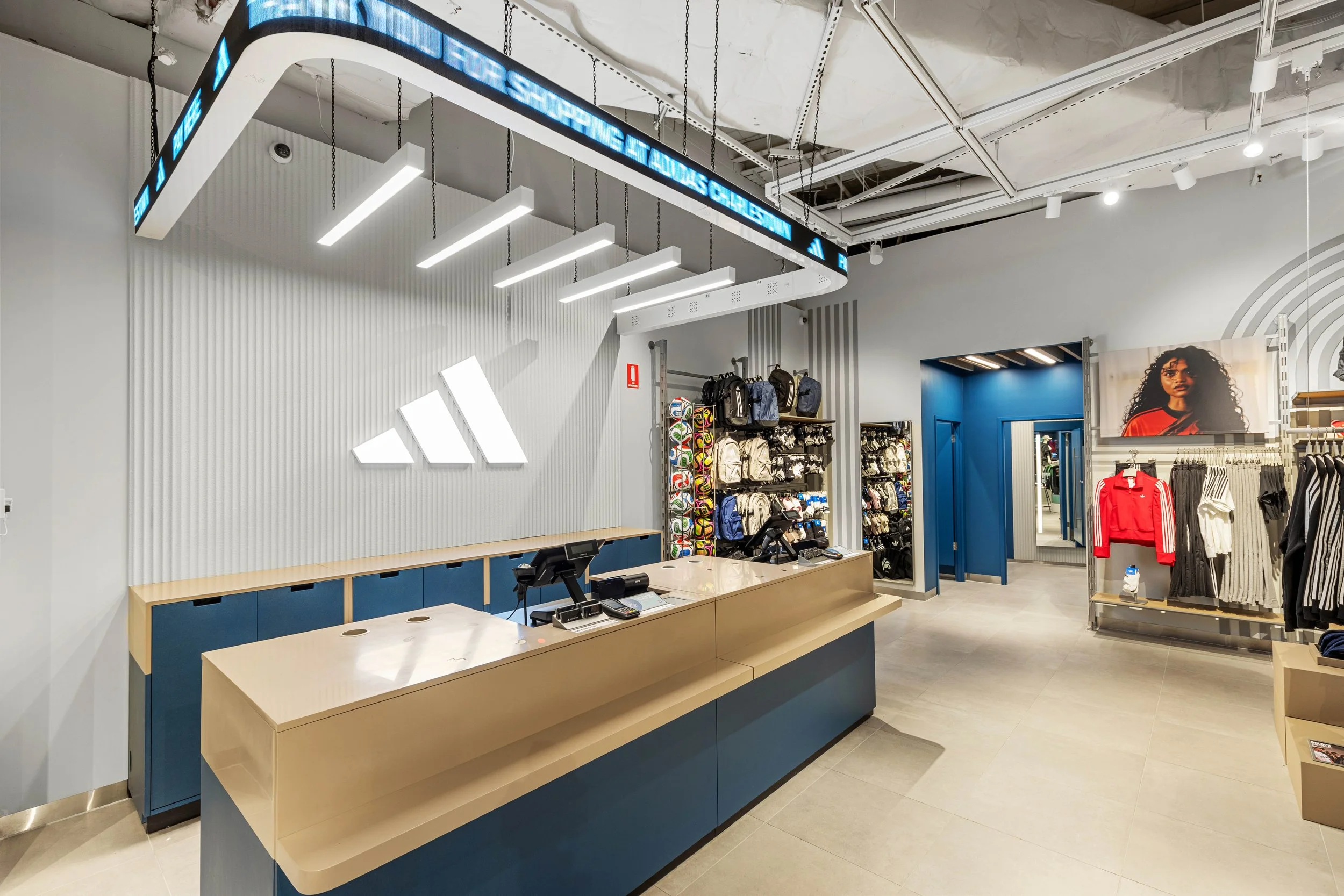 Interior of an Adidas store with a check-out counter, clothing and accessories on display, and a large Adidas logo on the wall.