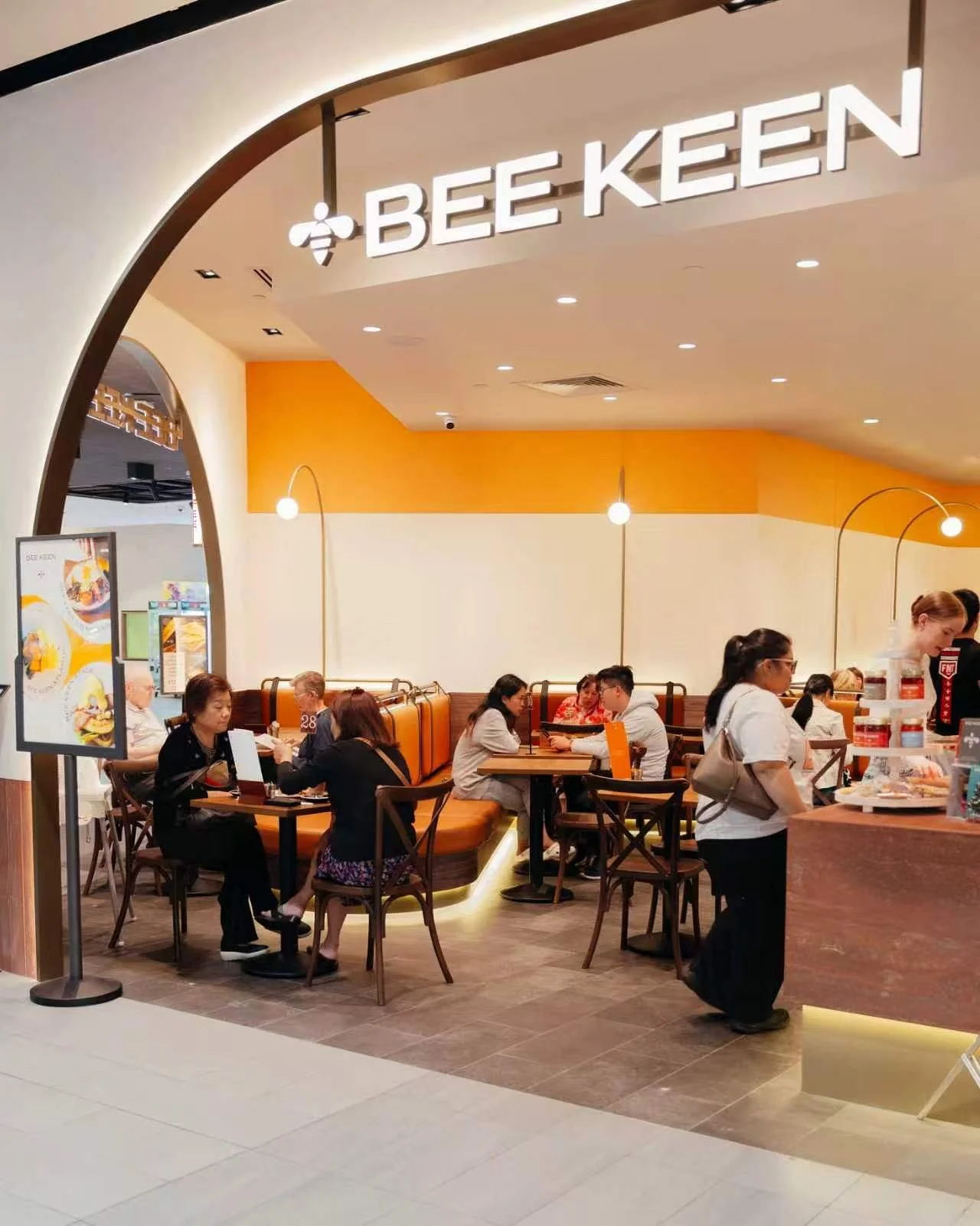 Inside a Bee Keen restaurant, customers are seated at tables, some eating and some talking, with a bright orange and white color scheme, modern lighting, and a menu display board near the entrance.