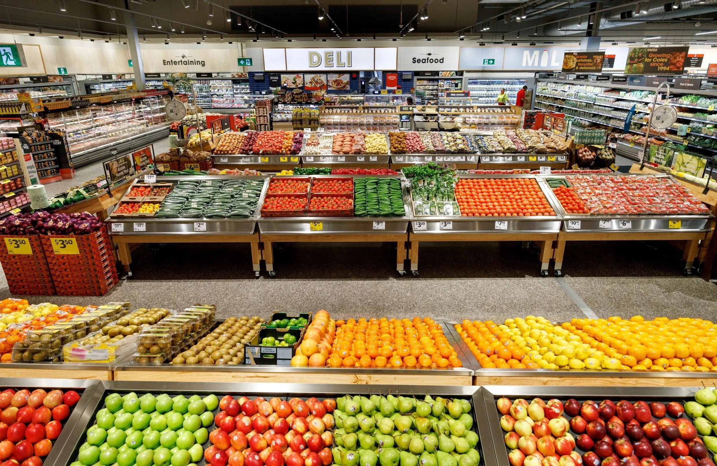 Interior of a grocery store with various fruits and vegetables displayed in bins and on shelves, including apples, tomatoes, and cucumbers, with signs and a deli section in the background.