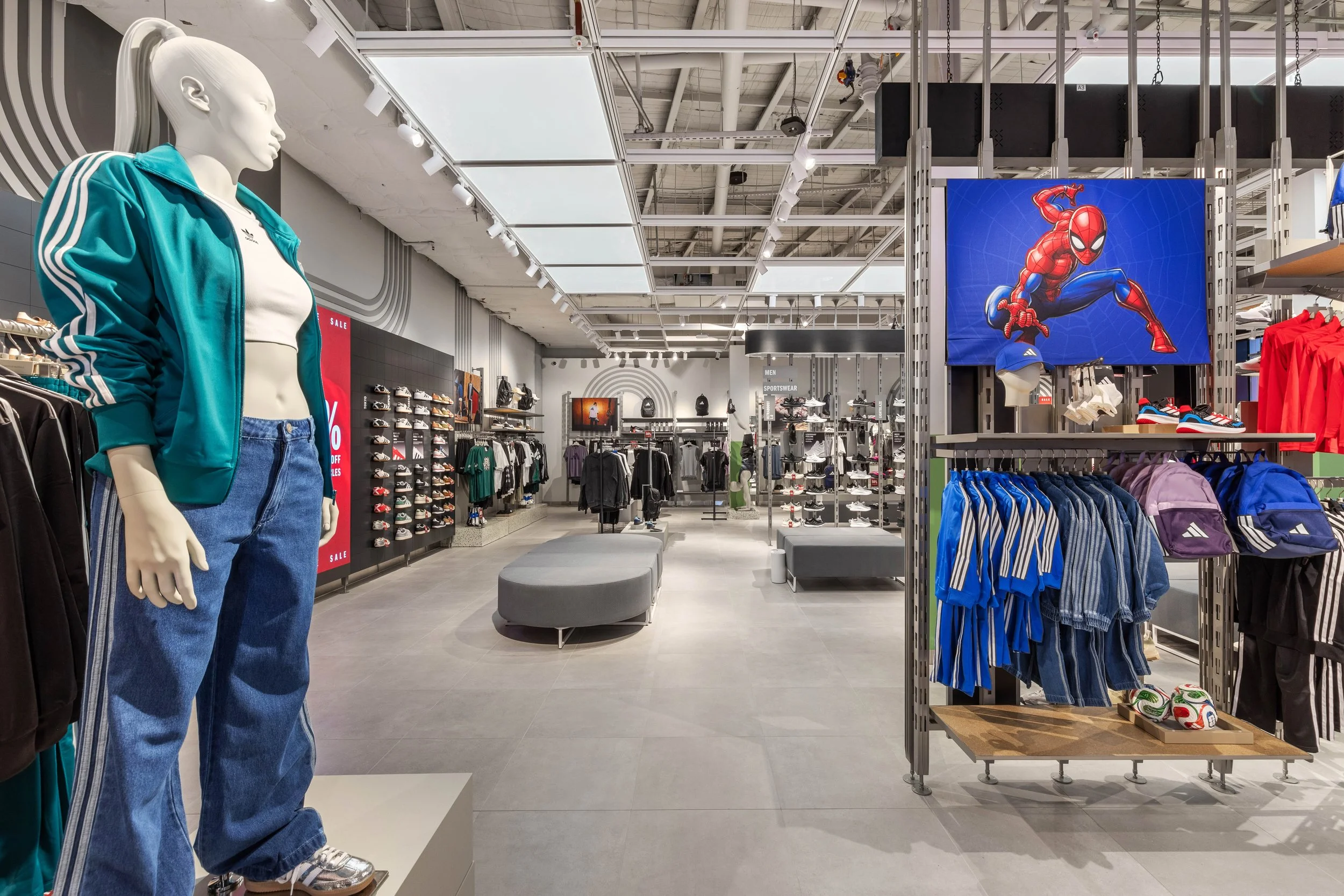 A mannequin dressed in a teal Adidas sports jacket and jeans in a retail store. The store has various sportswear and shoes on shelves and racks, with a large Spider-Man graphic on a display.