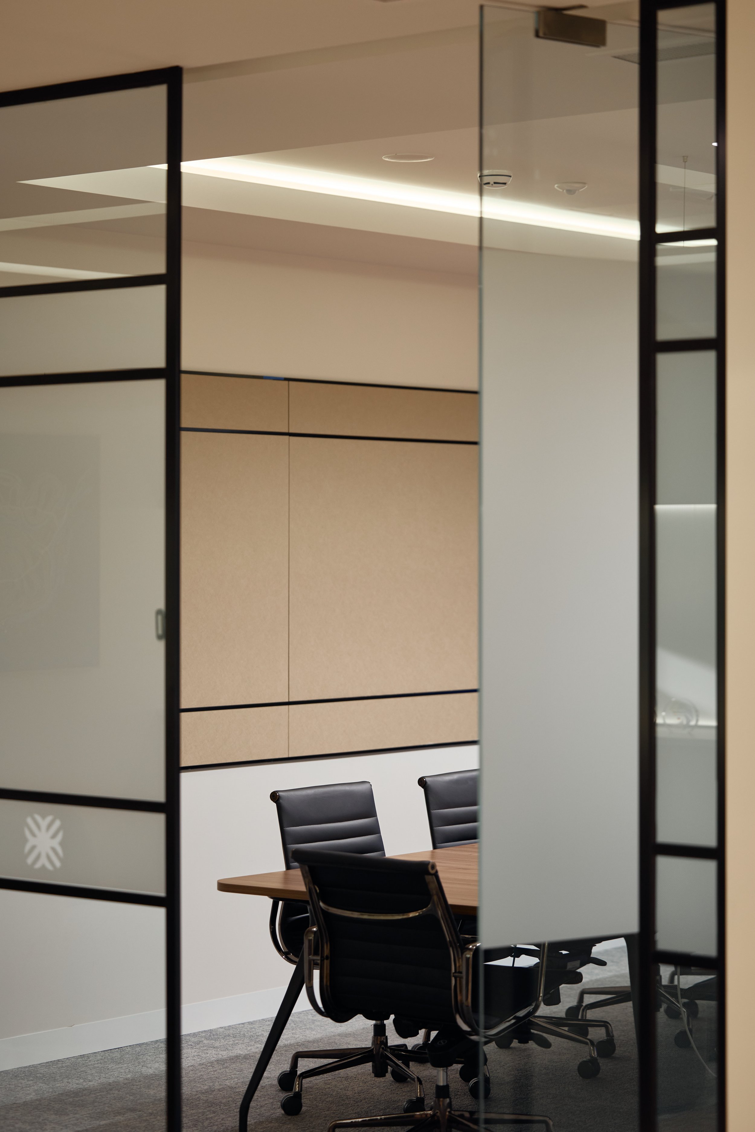 View of an empty modern conference room seen through glass doors with black frames. Inside, there is a wooden table, three black office chairs, and a beige wall with built-in cabinetry.
