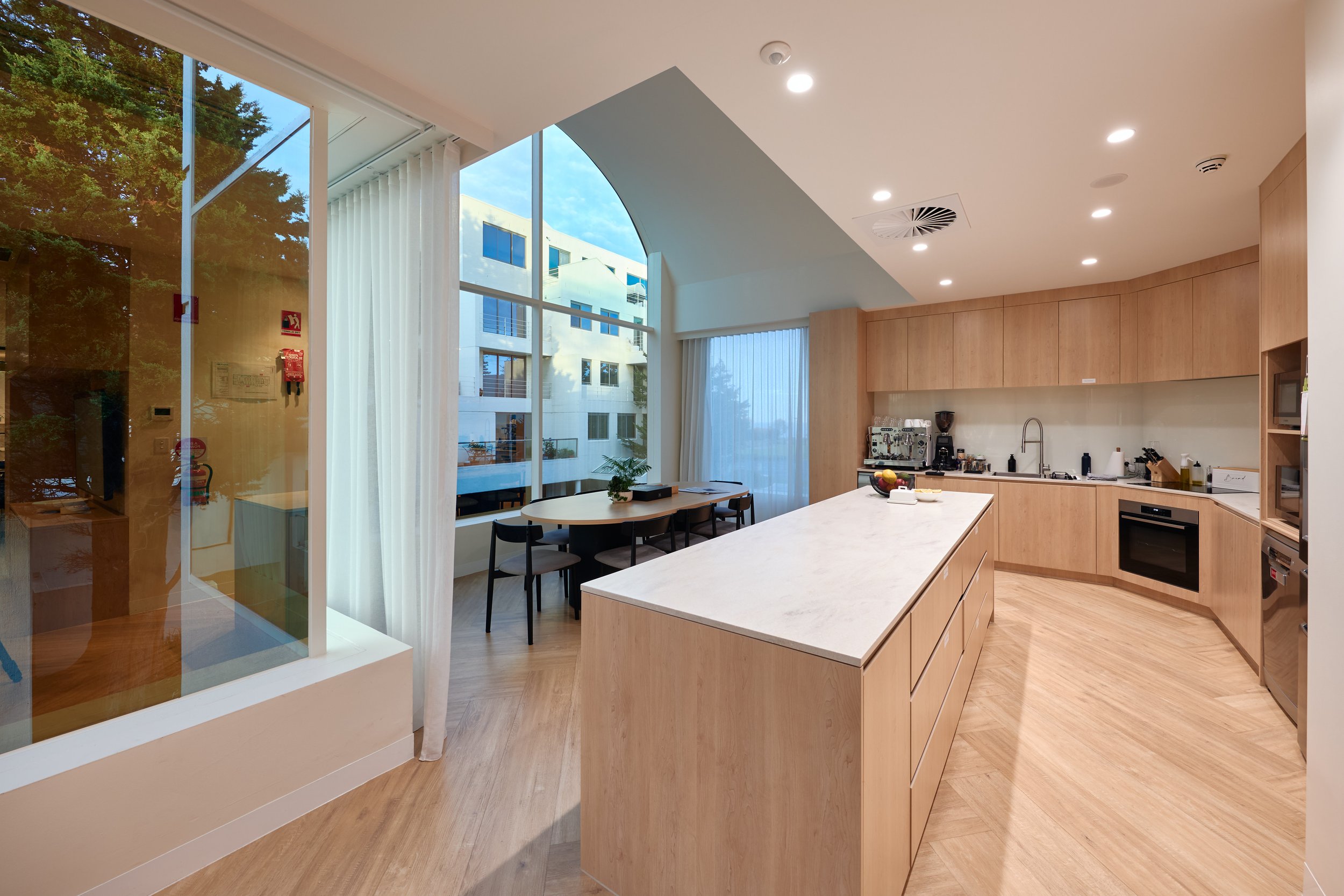 Interior view of a modern kitchen with white marble island, wooden cabinets, black chairs, and large windows showing neighboring buildings and trees outside.