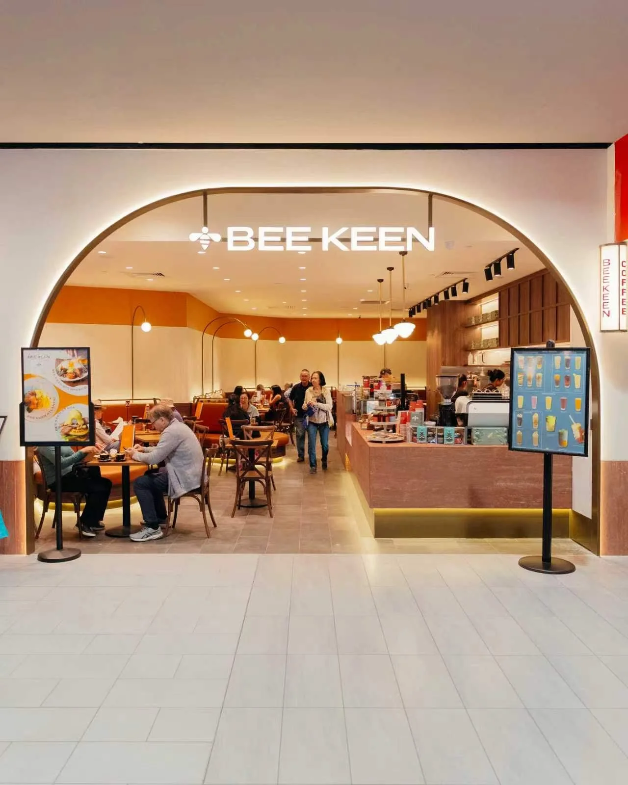 Interior of a Bee Keen cafe with customers sitting, a counter with staff, and digital menu boards.
