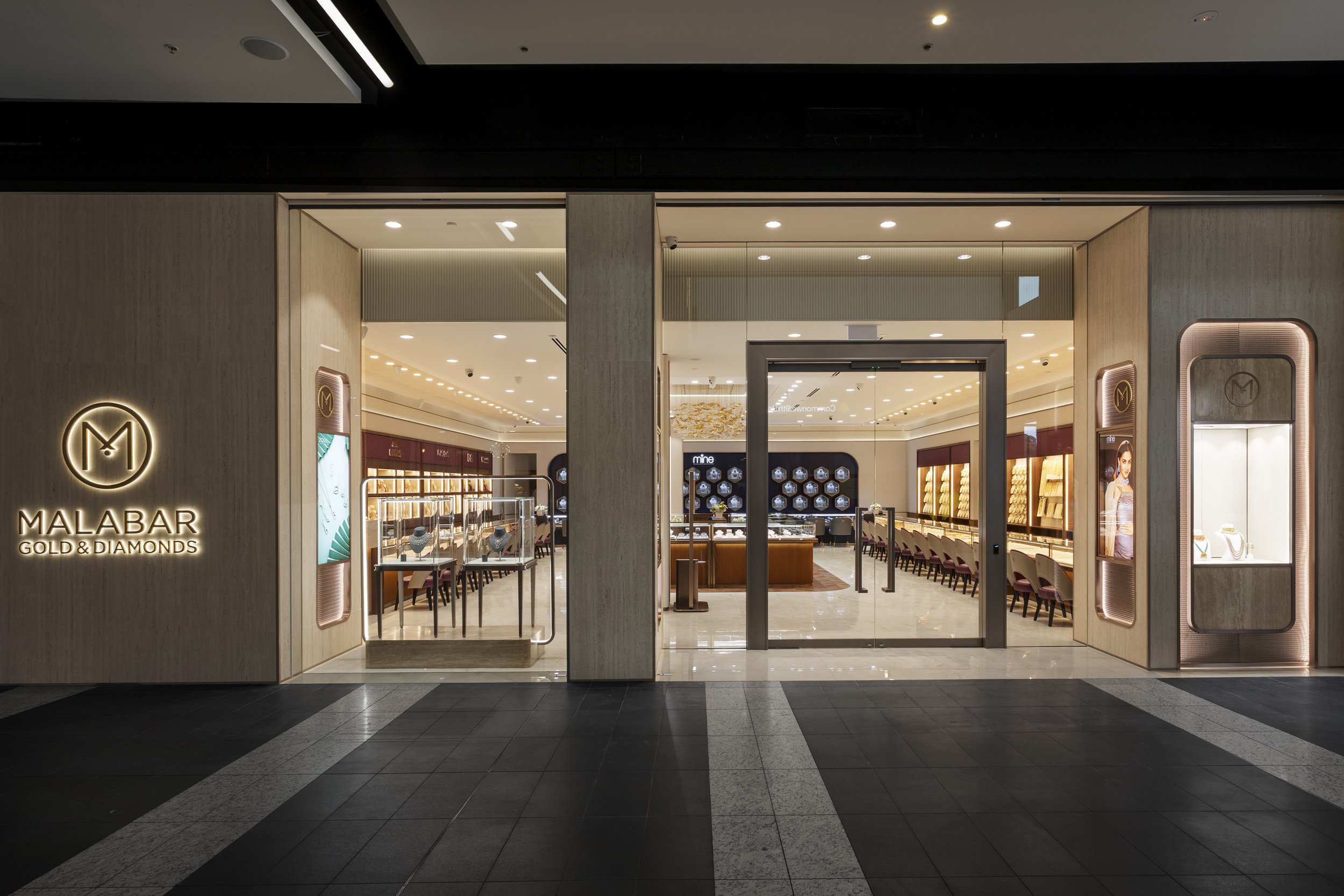 Entrance to a jewelry store named Malabar Gold & Diamonds, with a glass door and display window showcasing jewelry inside, illuminated with warm lighting, located in a shopping mall.