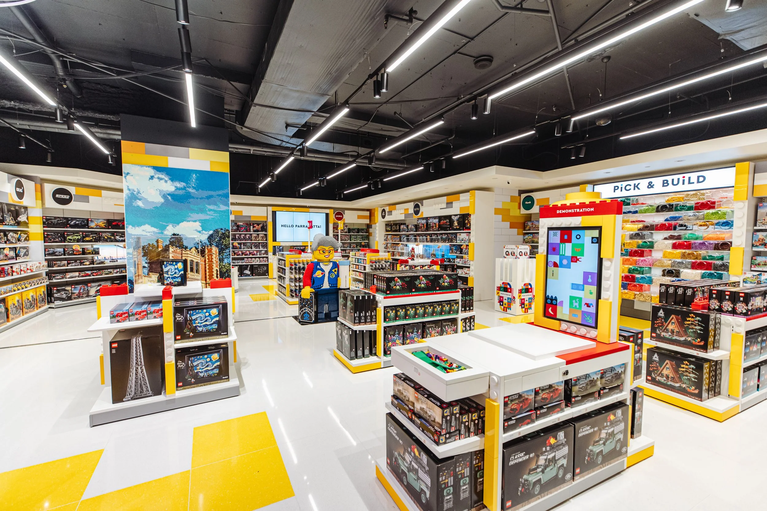 Interior of a LEGO store with shelves of LEGO sets, a large LEGO display of a person, and a digital screen, with bright lighting and yellow and white floor accents.