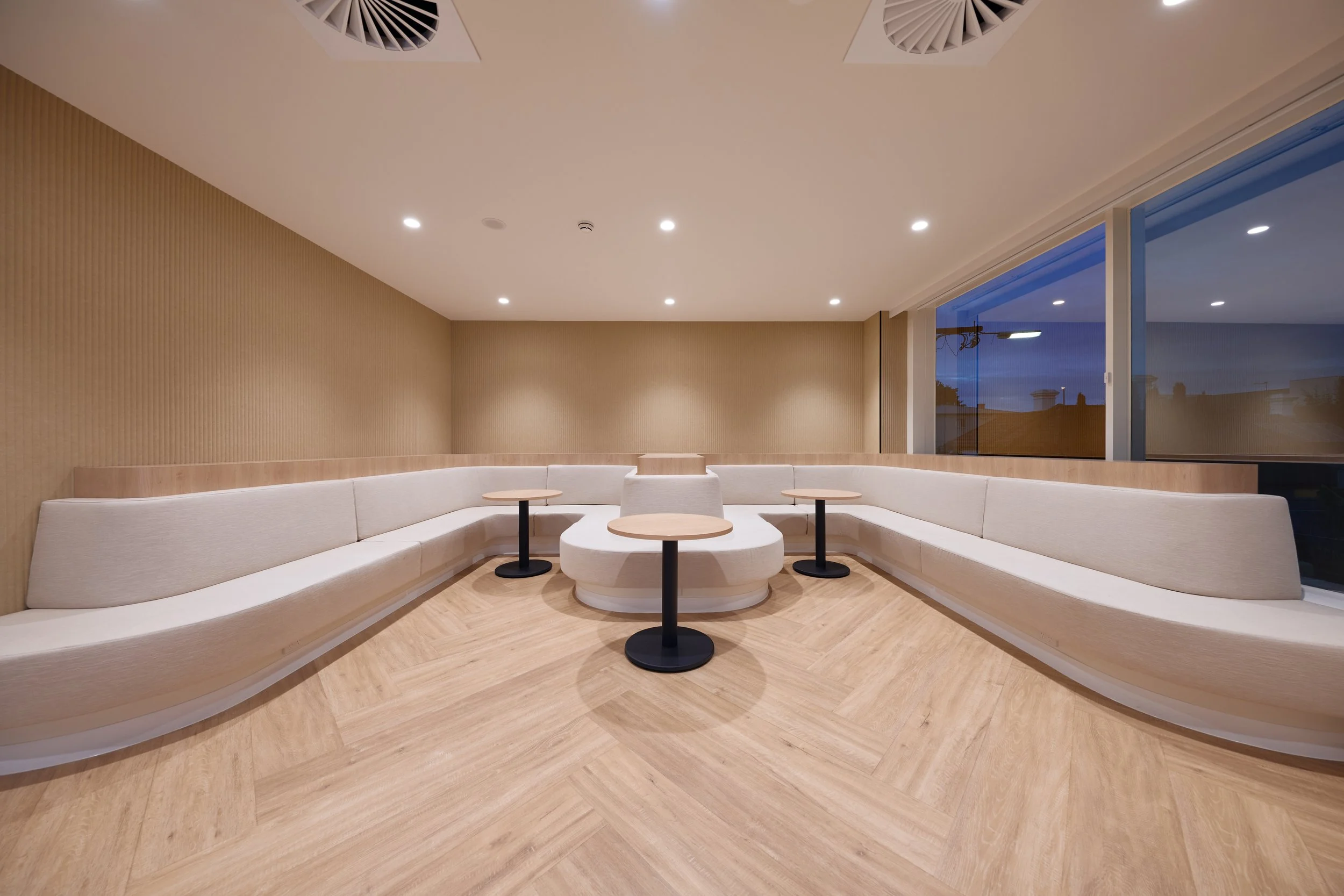 A modern lounge area with curved white seating, small round tables, large windows showing a nighttime cityscape, wood flooring, beige wall with textured paneling, and recessed ceiling lights.