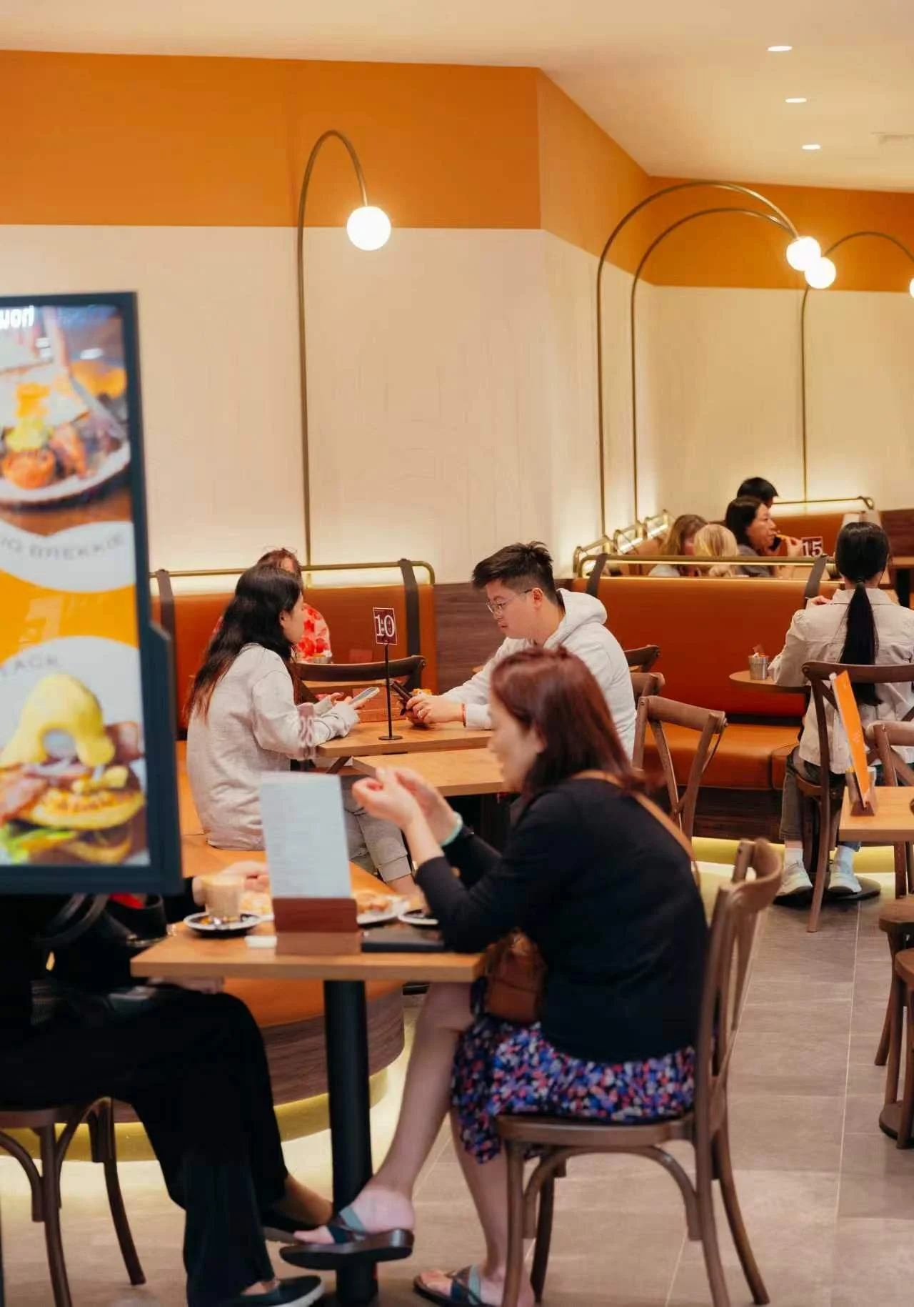 People dining at a restaurant with warm lighting, wooden furniture, and orange and beige walls, engaging in conversations and using their phones.