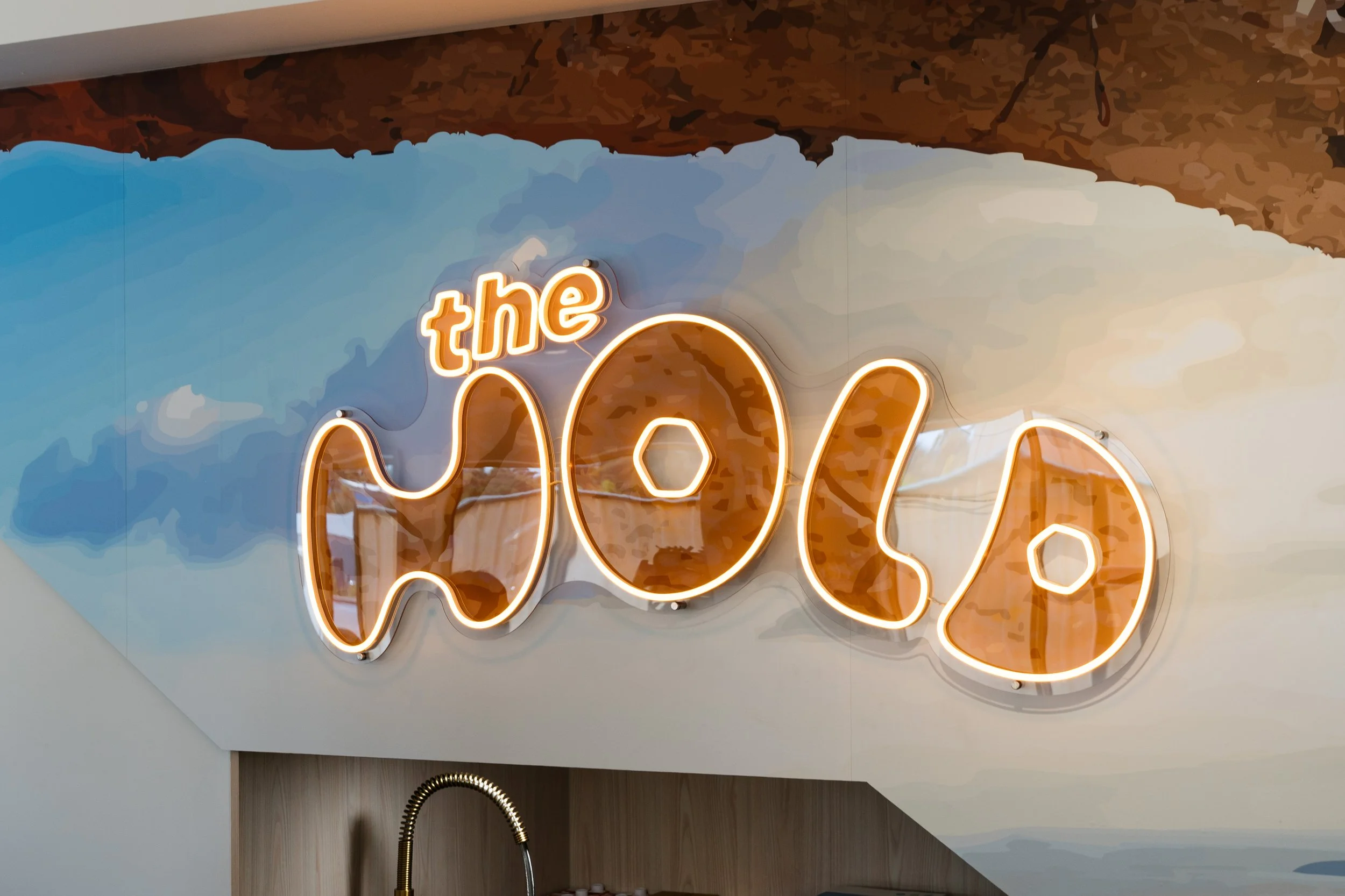 A neon sign with words in cursive lights, mounted on a wall with a blue sky and cloud artwork background.