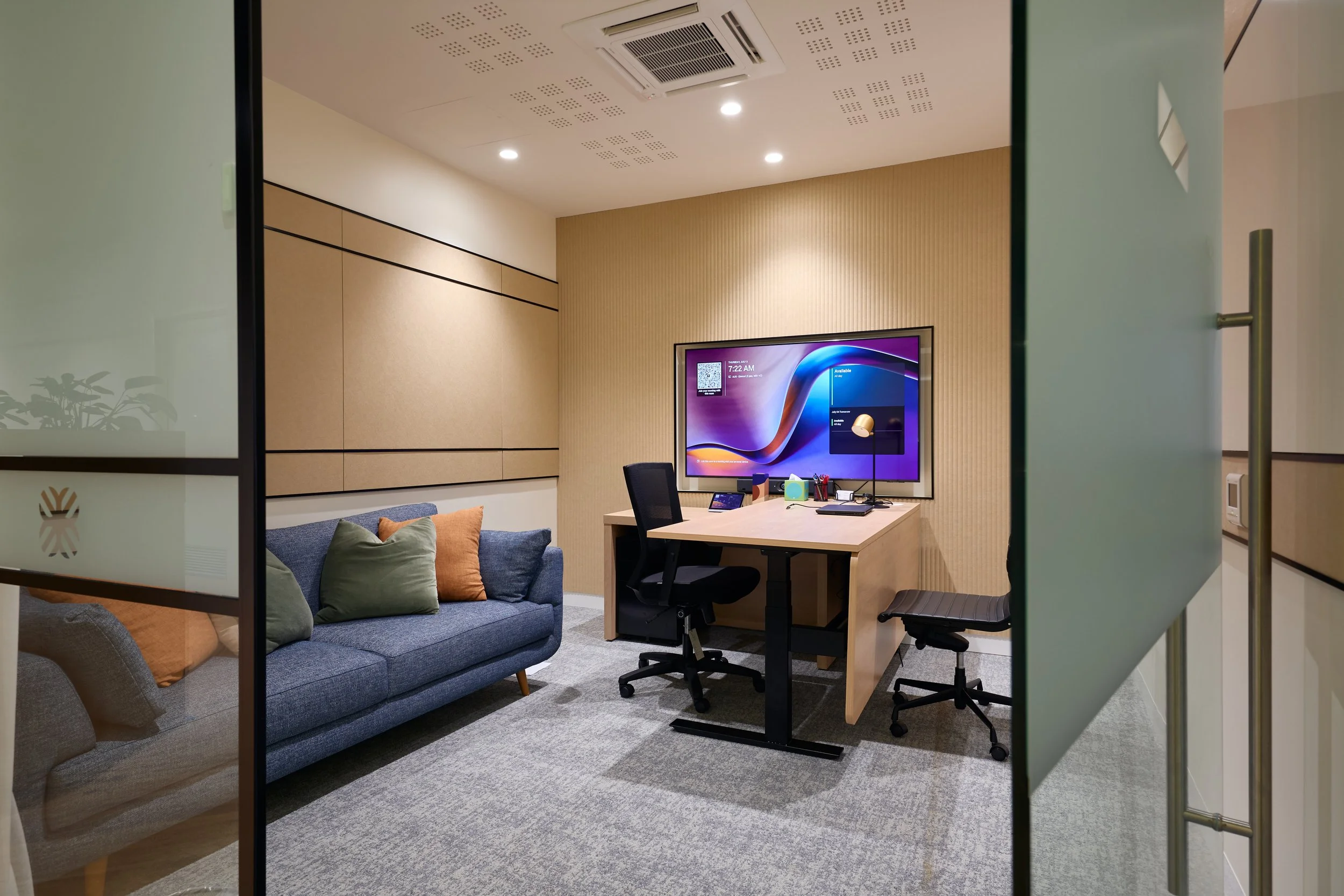 Office room viewed through partly open frosted glass doors with a black frame, featuring a gray sofa with multiple pillows, a wooden desk with a black office chair, a flat-screen TV mounted on a beige wall, and ceiling lights.