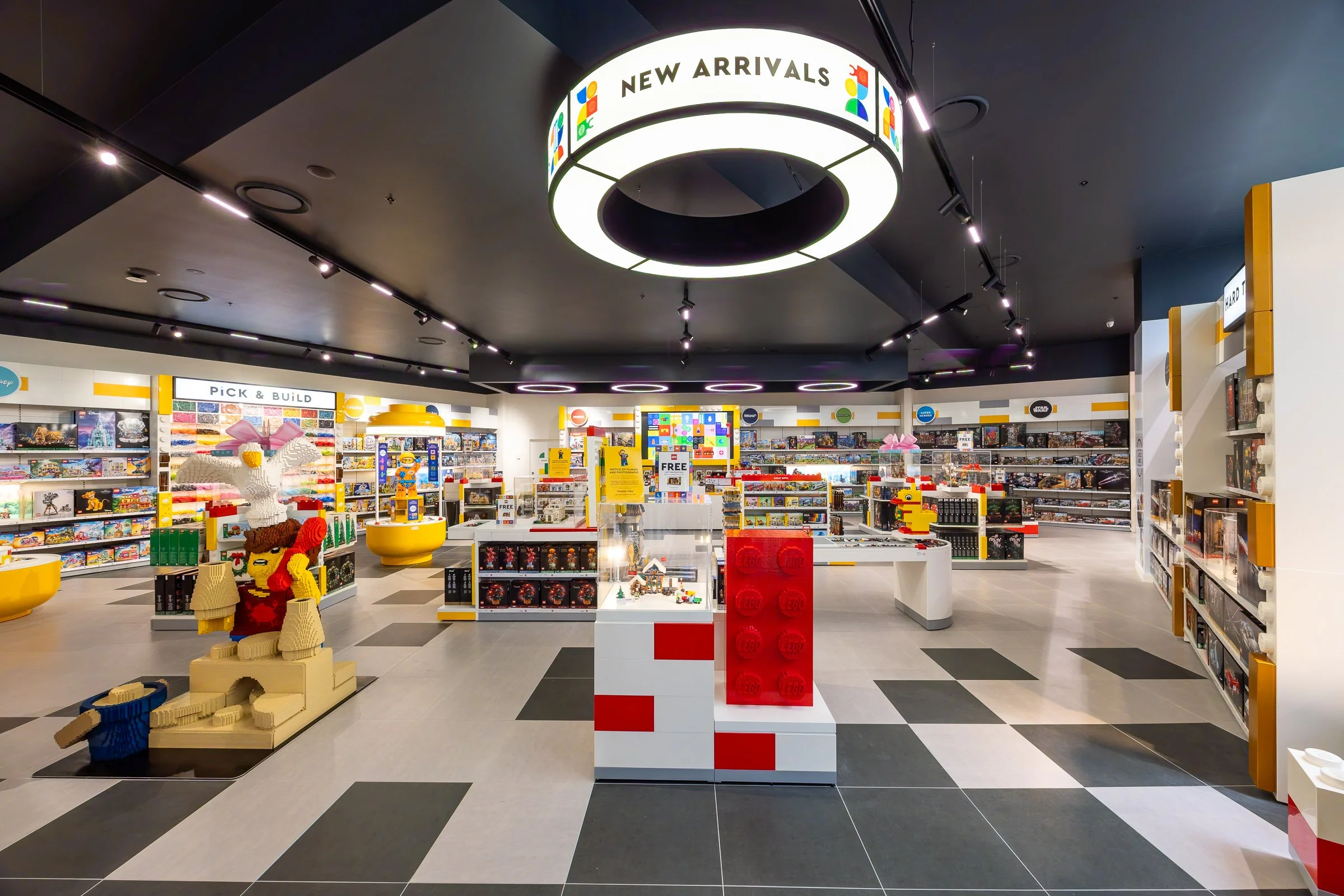 A retail toy store with shelves stocked with various toys and games. The store has a modern black and white checkered floor and brightly lit space.