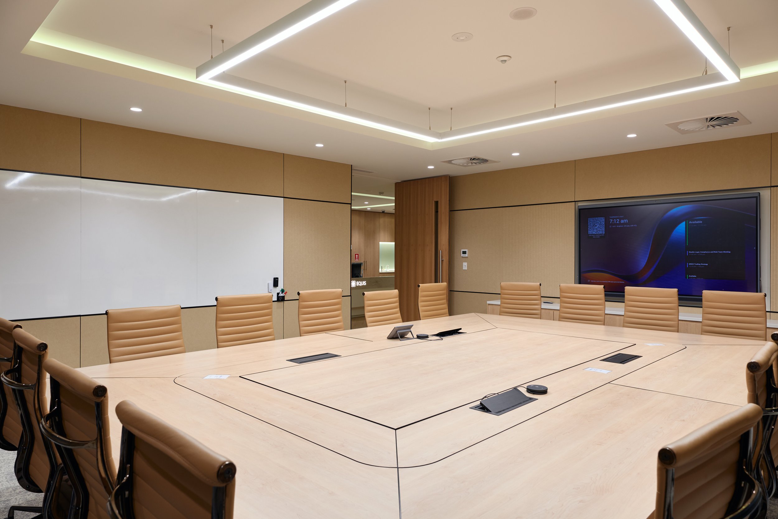 A modern conference room with a large light wood table surrounded by tan leather chairs, a whiteboard on one wall, a digital screen displaying a schedule, and ceiling LED lighting.