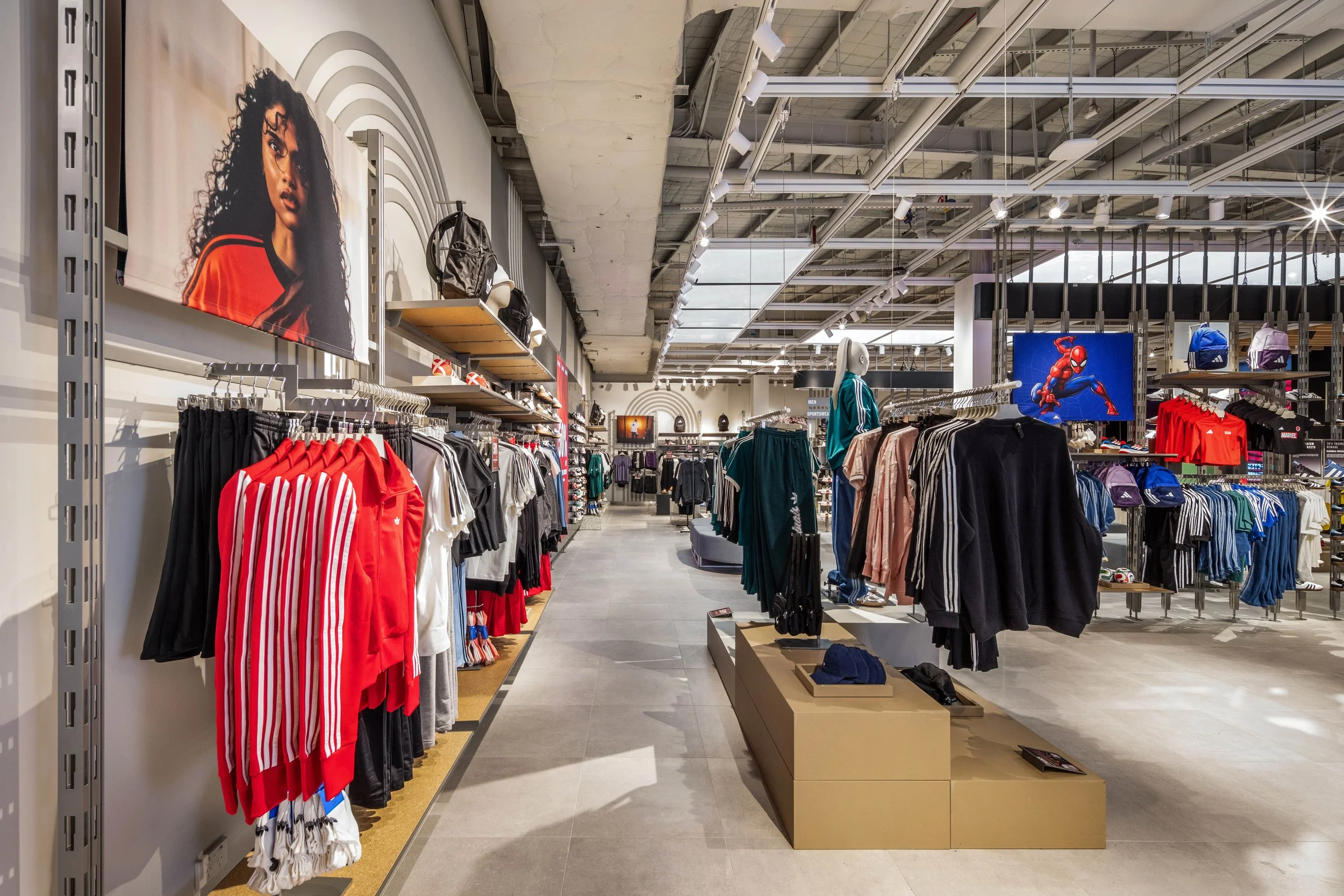 Interior of a modern retail store with athletic apparel, backpacks, and sports accessories, featuring posters of women and Spider-Man on the walls, with bright lighting and organized clothing displays.