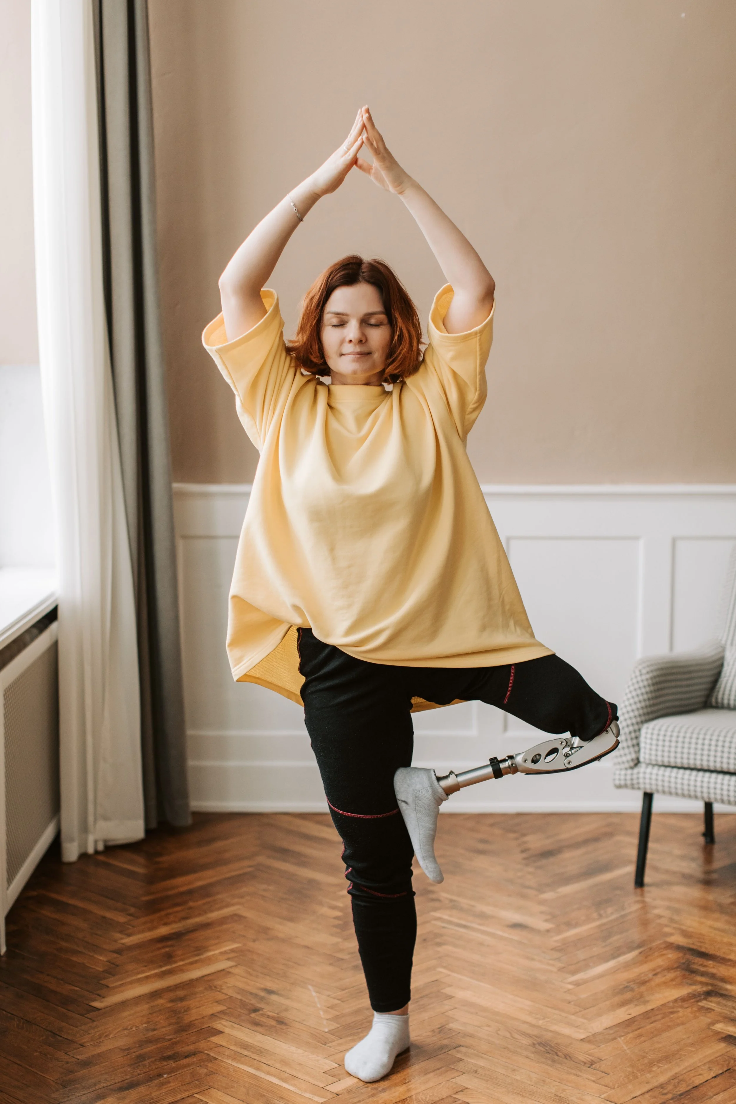A woman with a prosthetic leg practicing yoga indoors, standing on one leg with her arms raised above her head and hands pressed together, eyes closed, wearing a yellow T-shirt and black pants.