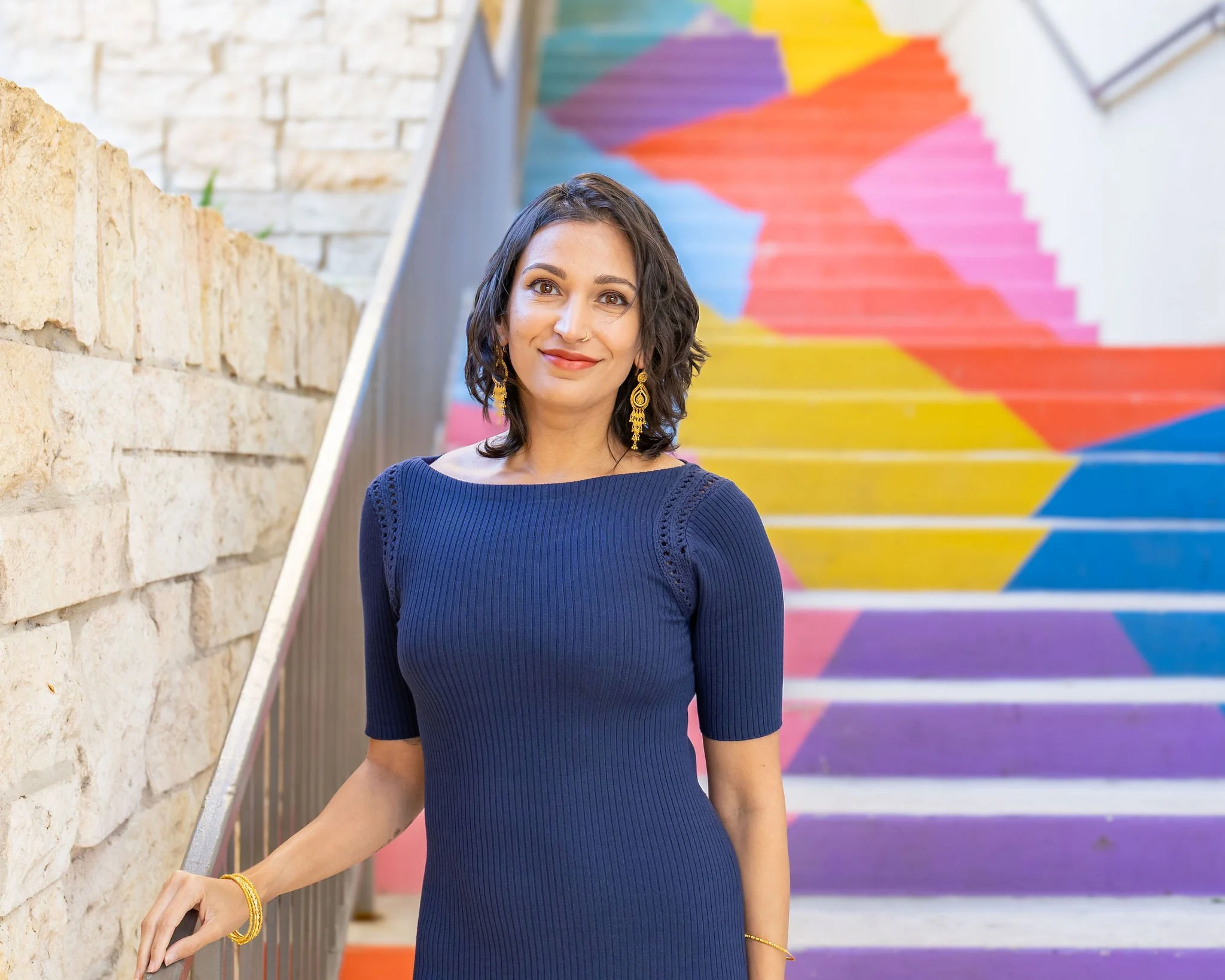 A woman with shoulder-length dark hair, wearing a blue dress and yellow earrings, standing in front of a colorful rainbow-painted staircase.