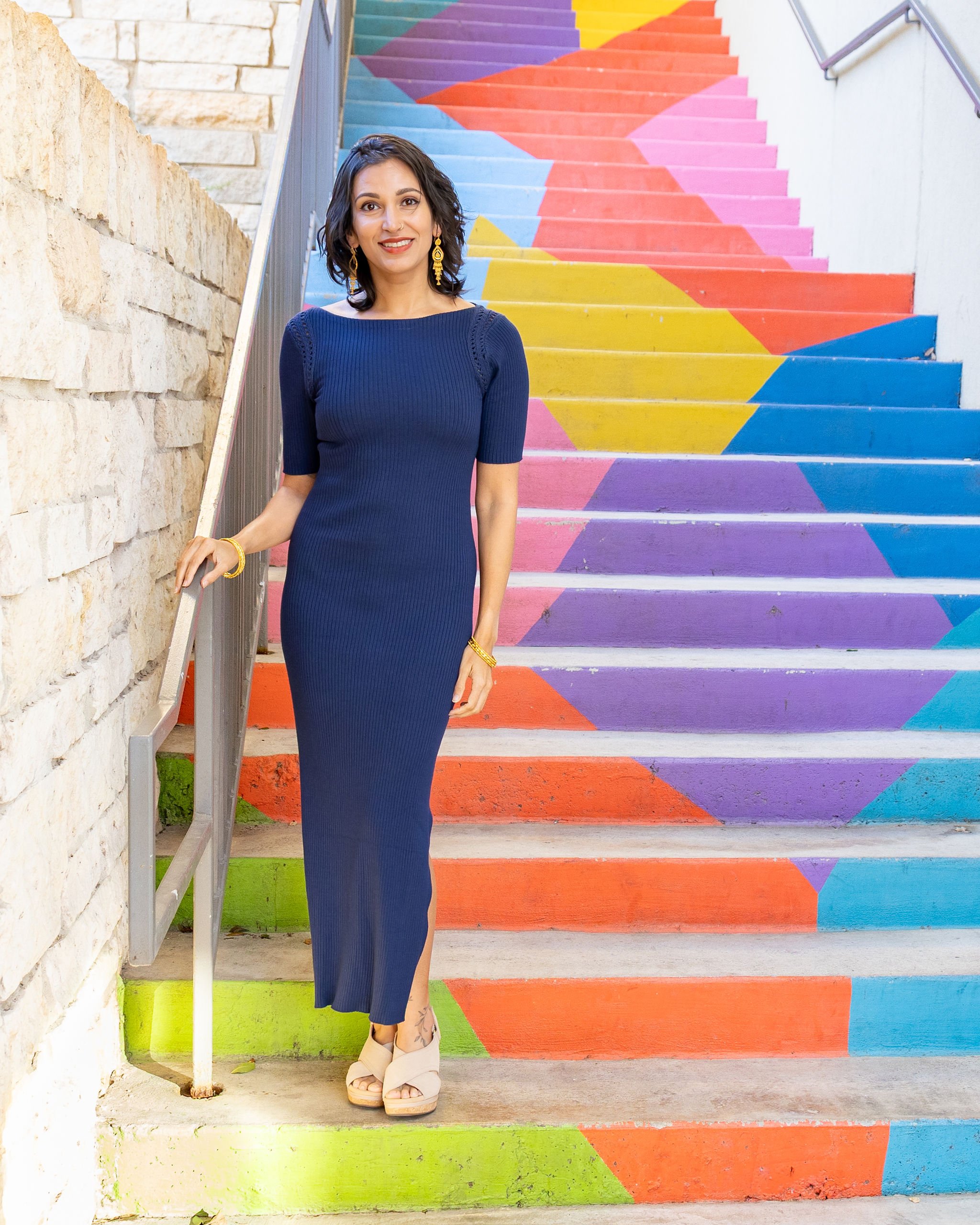 A woman wearing a navy blue dress and beige shoes standing on colorful painted stairs with geometric patterns in pink, yellow, blue, turquoise, and purple, next to a white brick wall and a metal handrail.
