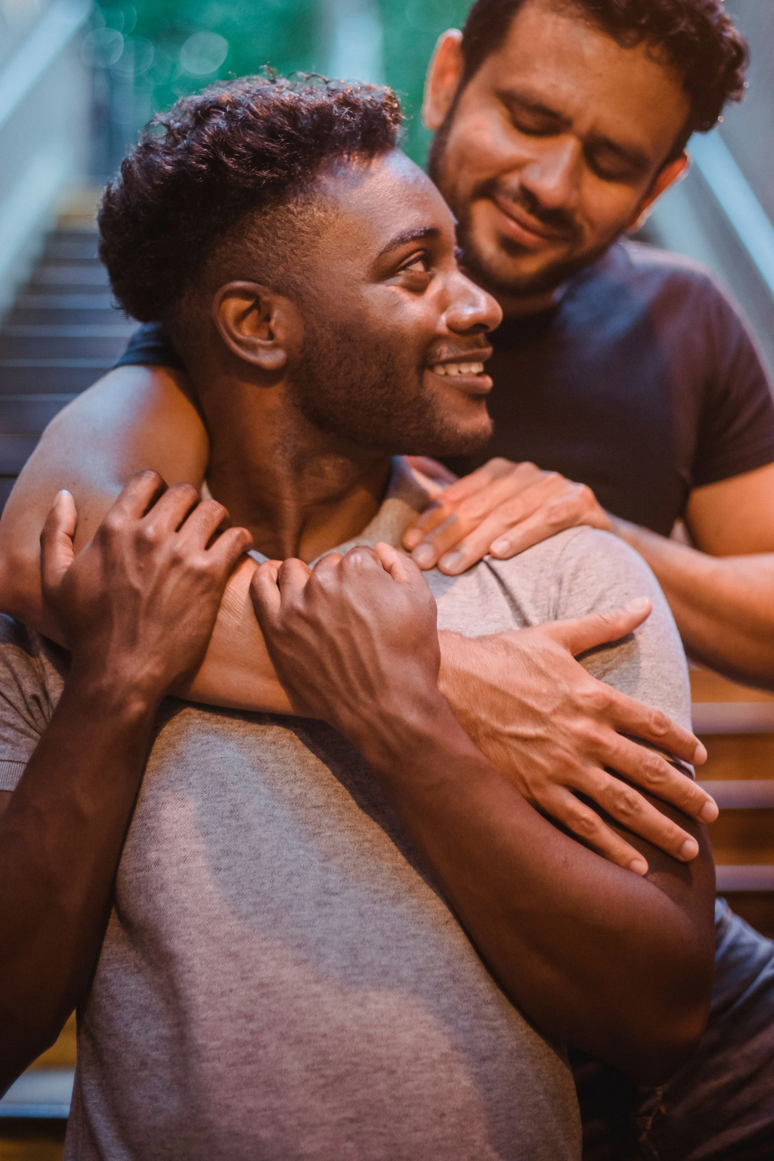 Two men hugging each other with smiles, one with dark curly hair and the other with short black hair and glasses, indoors near a staircase.