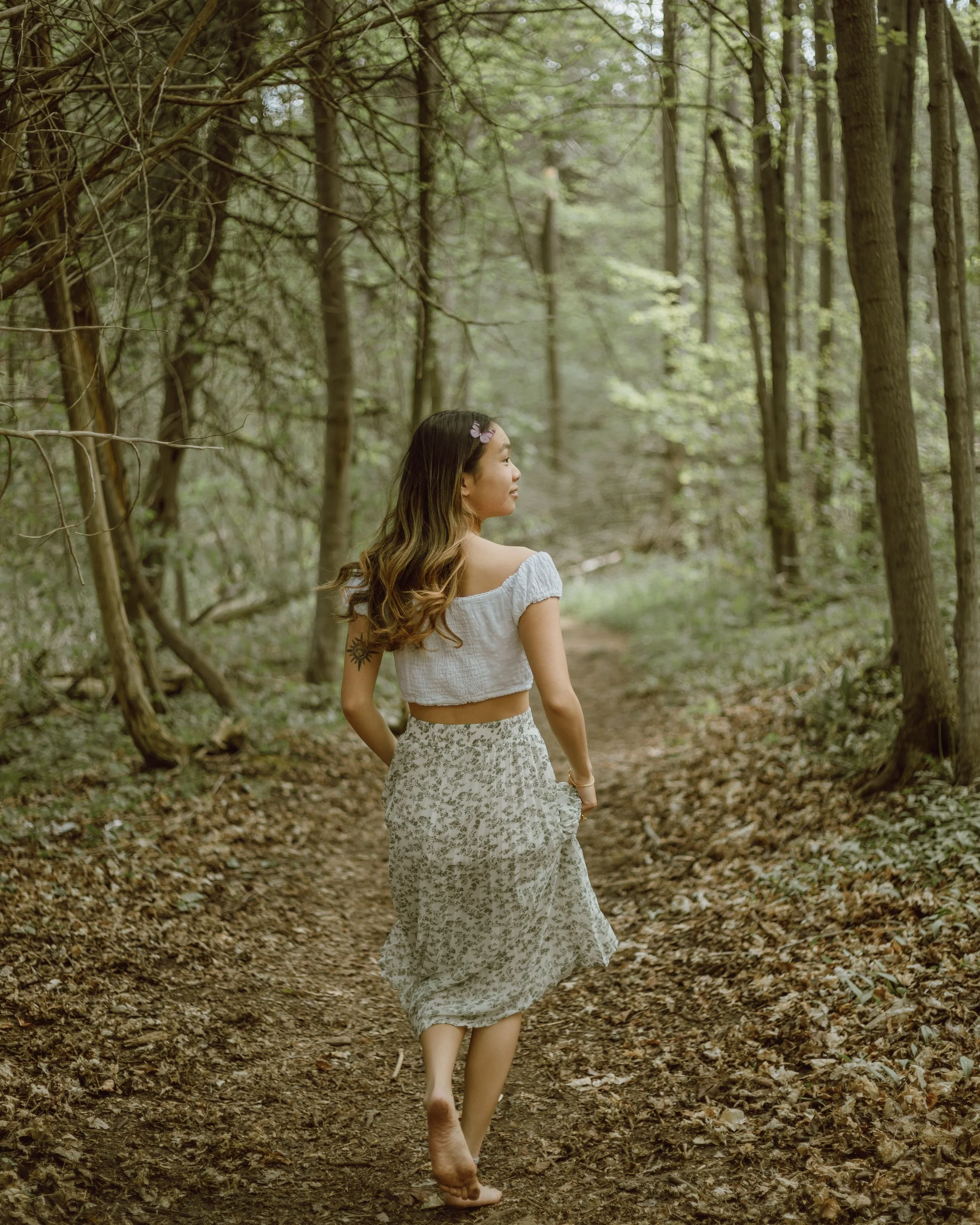A young woman with long, wavy hair and a butterfly hair clip, wearing a white off-the-shoulder crop top and a long floral skirt, walking barefoot on a dirt path in a lush, green forest.