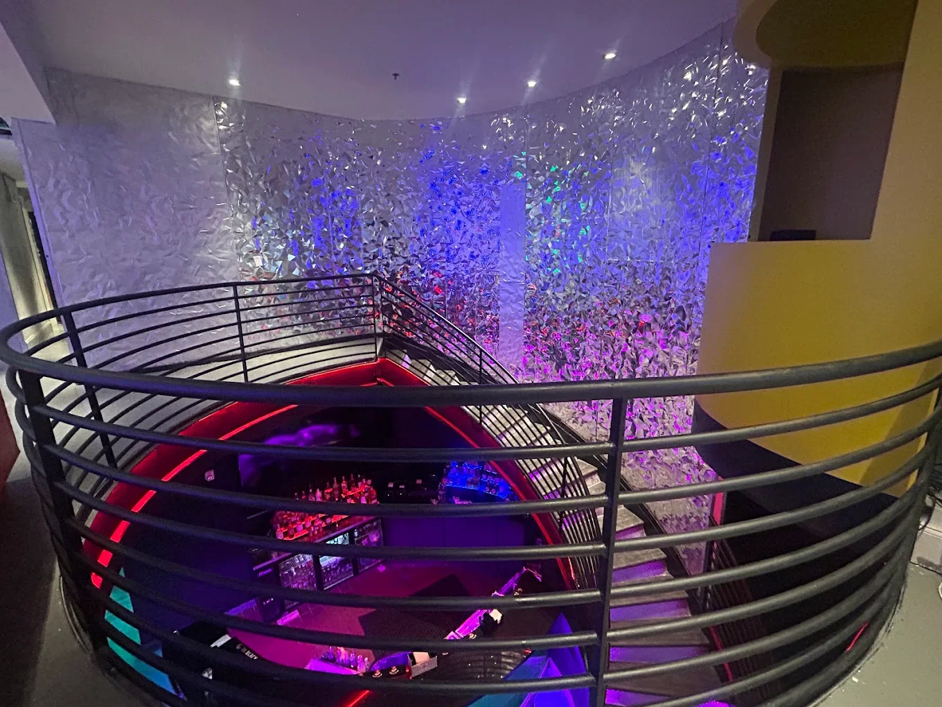 A spiral staircase with metal railing leading down to a bar area illuminated with purple and red lighting. The background features a textured wall with reflective, crinkled metallic surface and colorful lights.