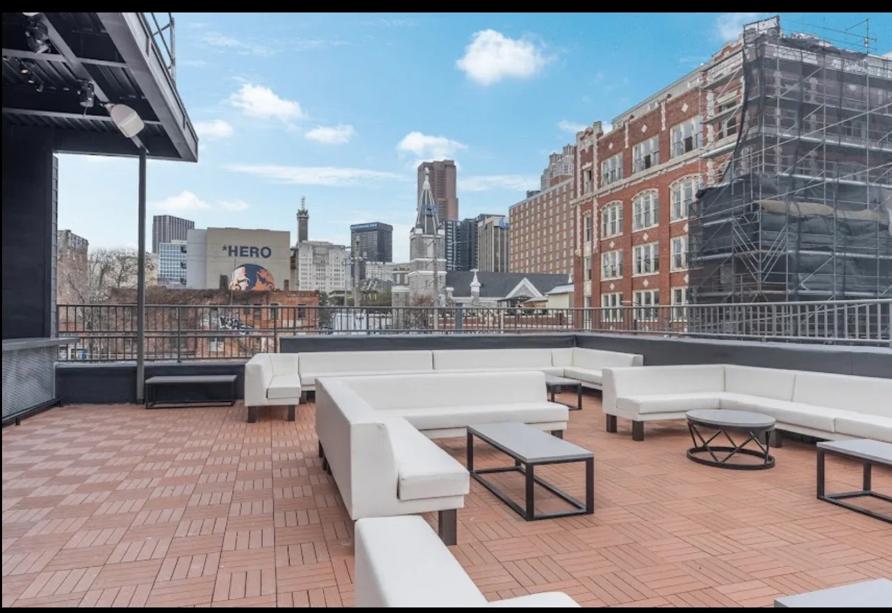 Rooftop terrace with white outdoor furniture, few tables, and city skyline in the background, including buildings and a billboard that reads "HERO."