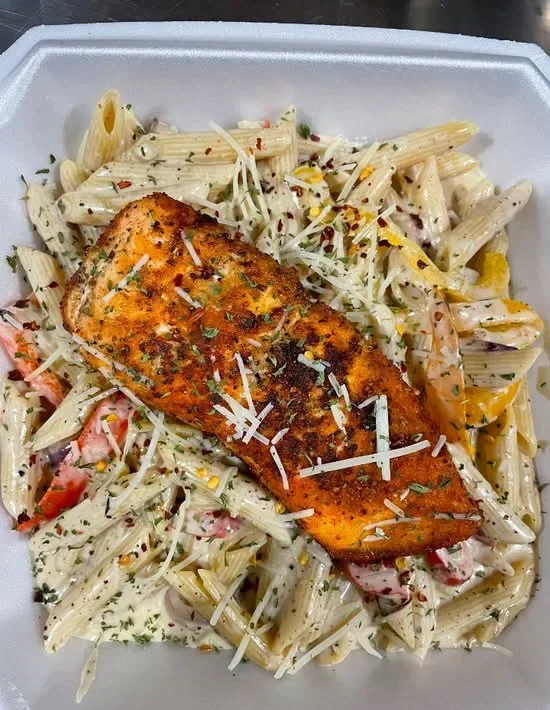Grilled salmon fillet on a bed of penne pasta with creamy sauce, broccoli, and shredded cheese, garnished with herbs and spice flakes.