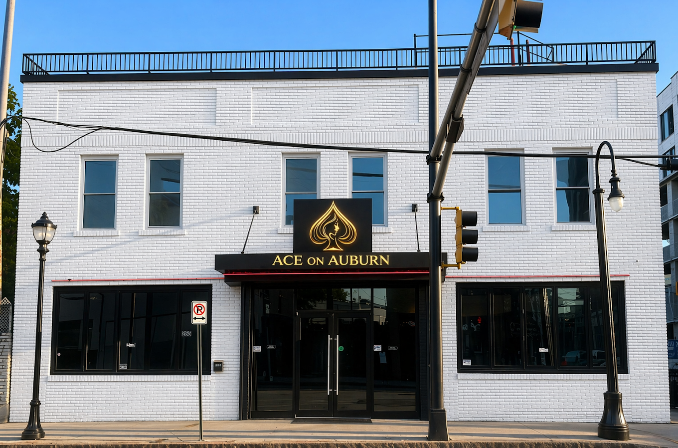 Exterior of a white brick building with large black-framed windows and a black sign that says "ACE on AUBURN" with a gold logo of a spade and a face silhouette above it. There are street lamps, traffic lights, and a no parking sign in front of the bu