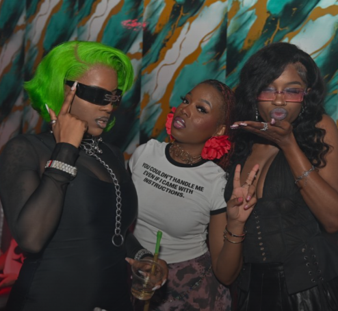 Three women posing at a party with colorful abstract background. The woman on the left has vivid green hair, stunt glasses, and is holding a drink. The woman in the middle has reddish-brown hair, tattoos on her neck, and wears a white shirt. The woman on the right has black curly hair, sunglasses, and is making a peace sign.