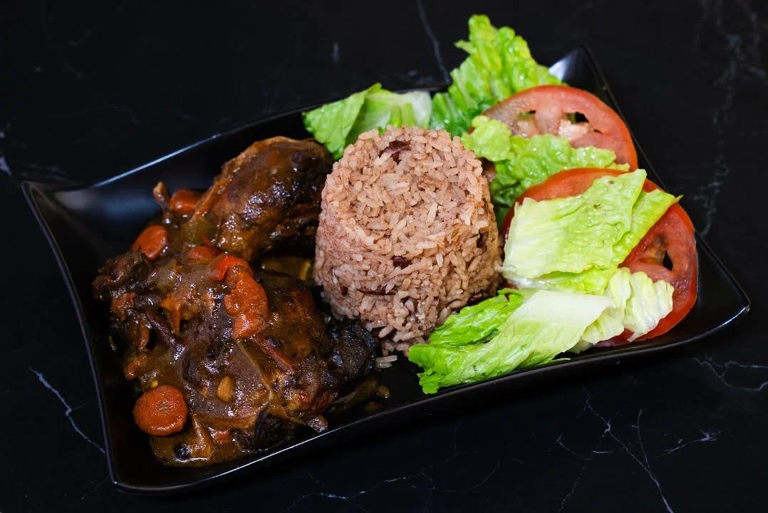 Steamed brown rice, lettuce, and tomato slices with a meat stew featuring vegetables like carrots in a dark sauce on a black plate.