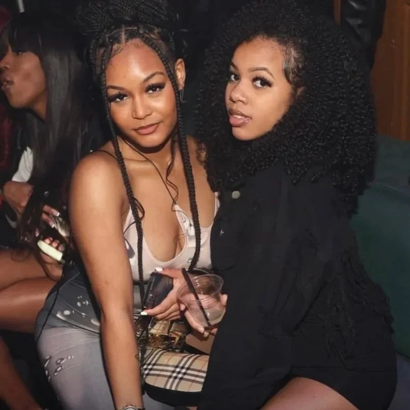 Two young women with curly and braided hairstyles are sitting close together at a social event, smiling and holding drinks.