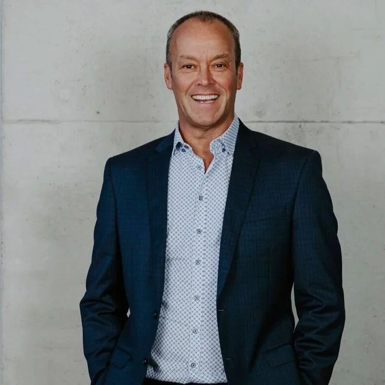 Smiling middle-aged man in a blue suit and white patterned shirt standing against a plain wall.