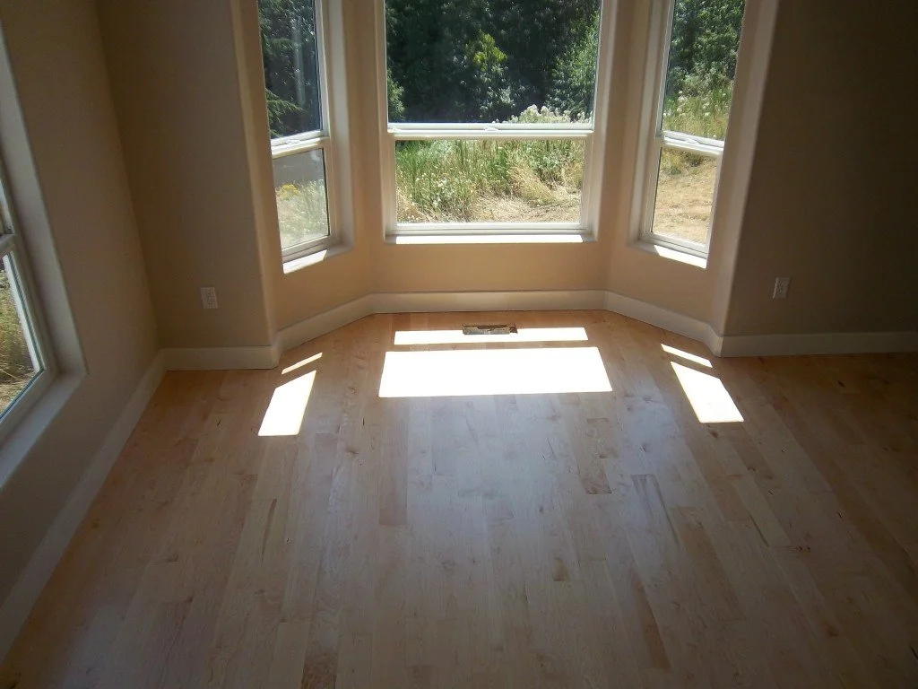 Empty room with large windows letting in sunlight, revealing light wood flooring and a view of greenery outside.