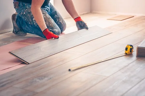 Person installing hardwood flooring, kneeling on the floor, wearing gloves, with tools nearby.