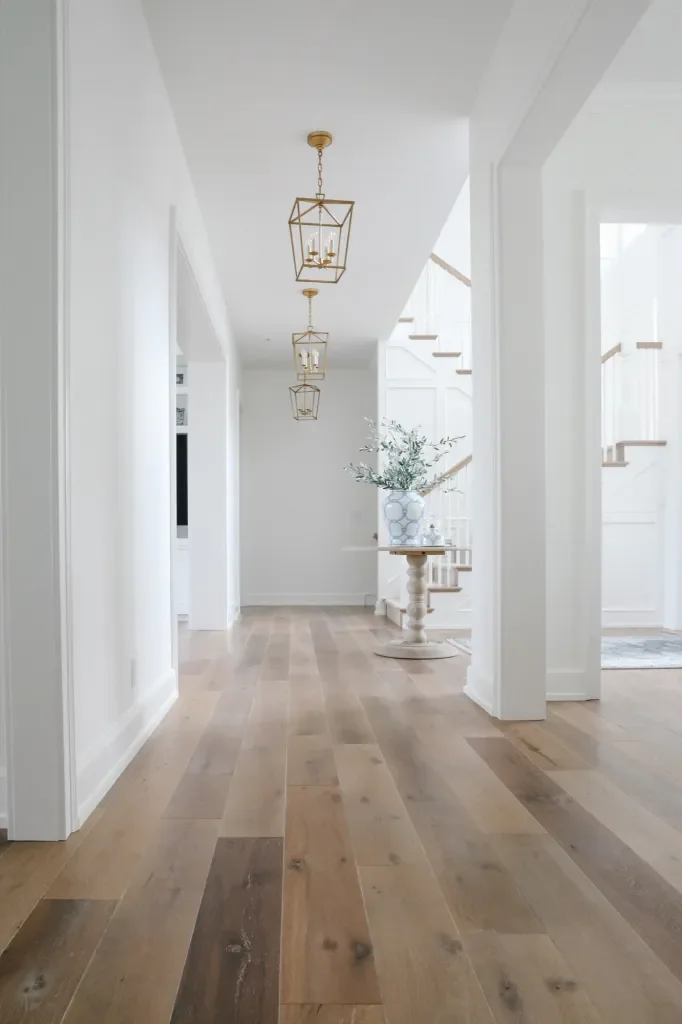 Bright white hallway with gold lantern pendant lights, wooden flooring, and a staircase with a potted plant on a round table near the stairs.
