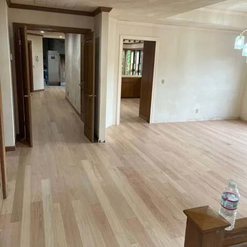 Empty living room with light wood floors and white walls, visible doorways, and a water bottle on a small table.
