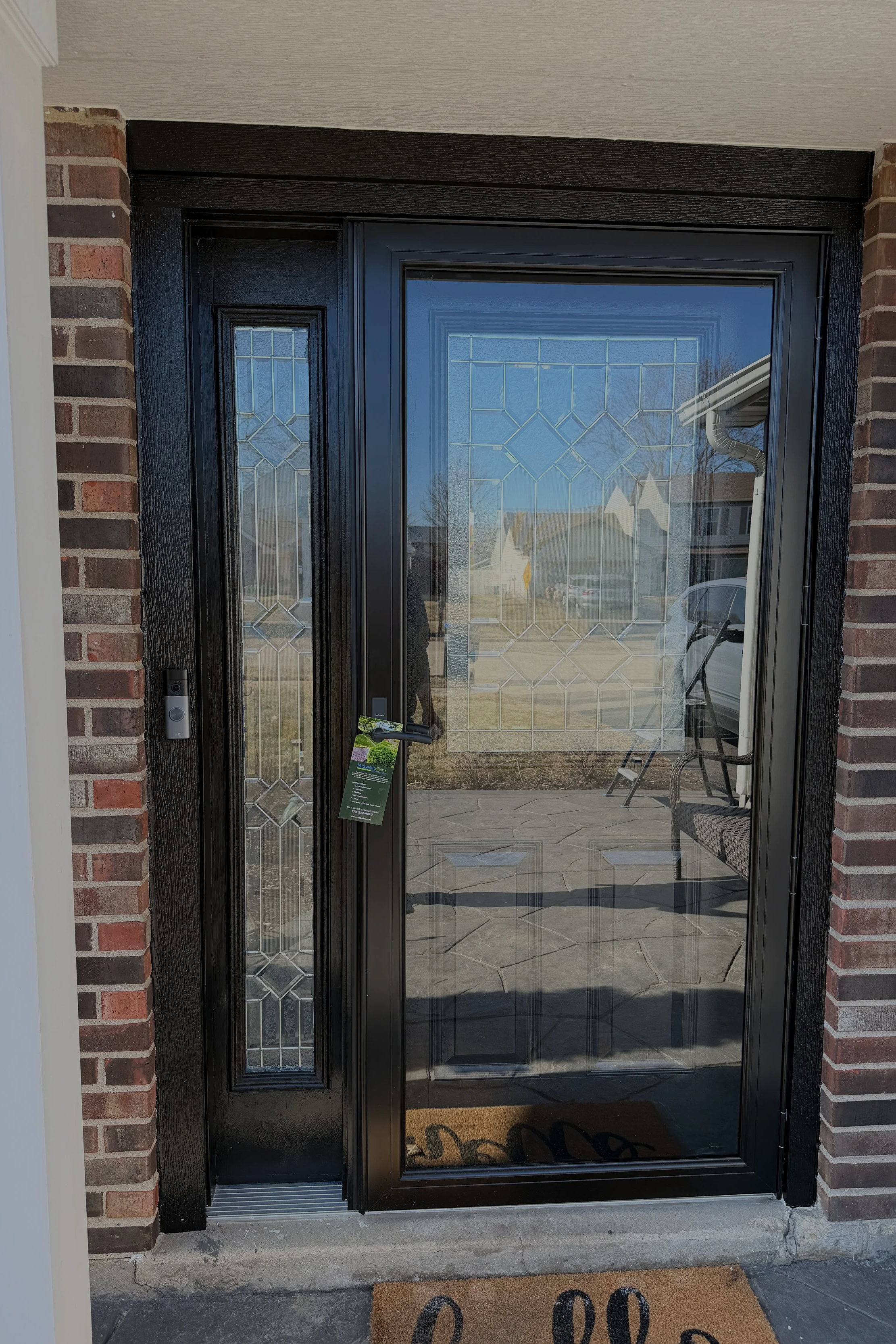 Front door with black frame and decorative glass panel, brick wall siding, concrete step, and 'hello' doormat.