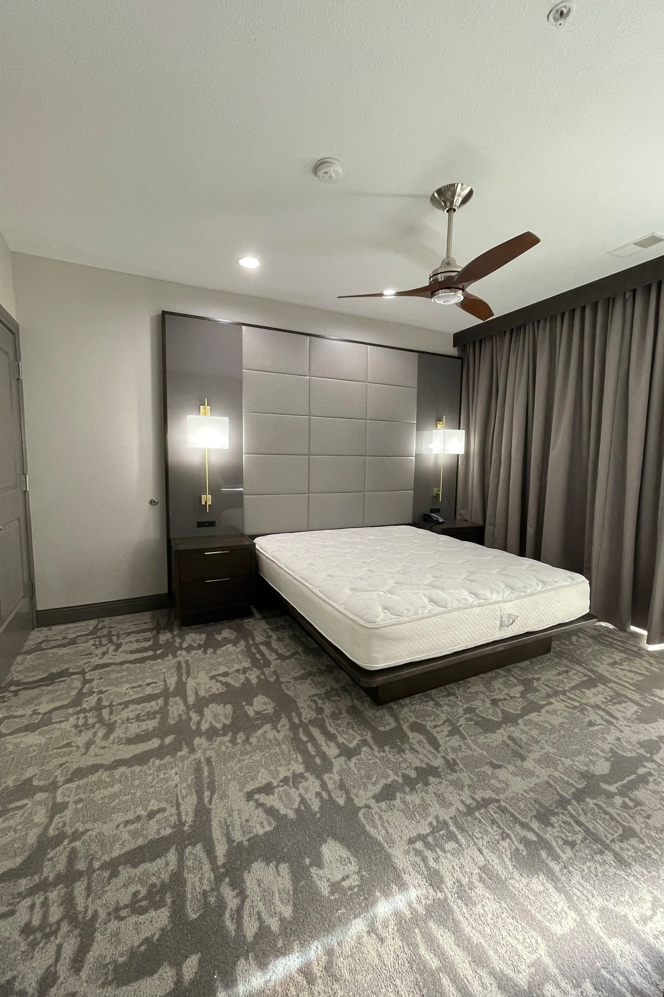 room with a bed, headboard, two nightstands, wall-mounted lights, a ceiling fan, and floor-length curtains.