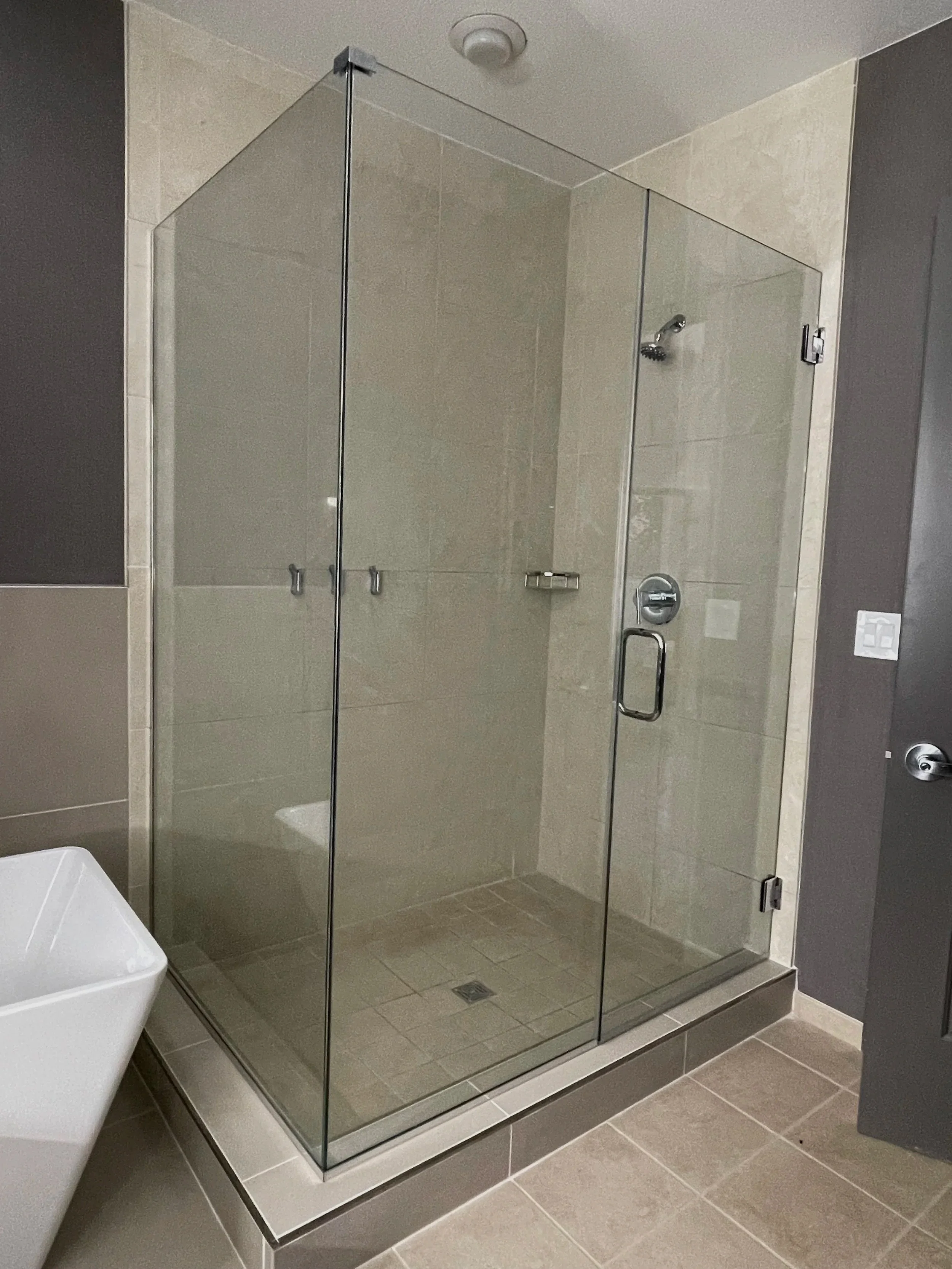 A modern shower with clear glass walls, beige tiled walls and floor, a silver showerhead inside, and a silver door handle.