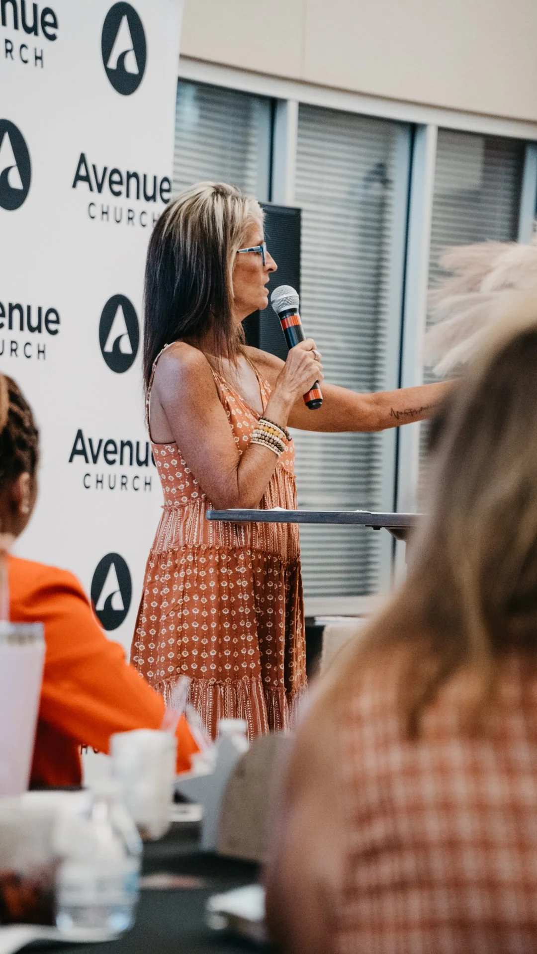 Woman in an orange dress speaking into a microphone at a church event with a white backdrop that has the 'Avenue Church' logo.