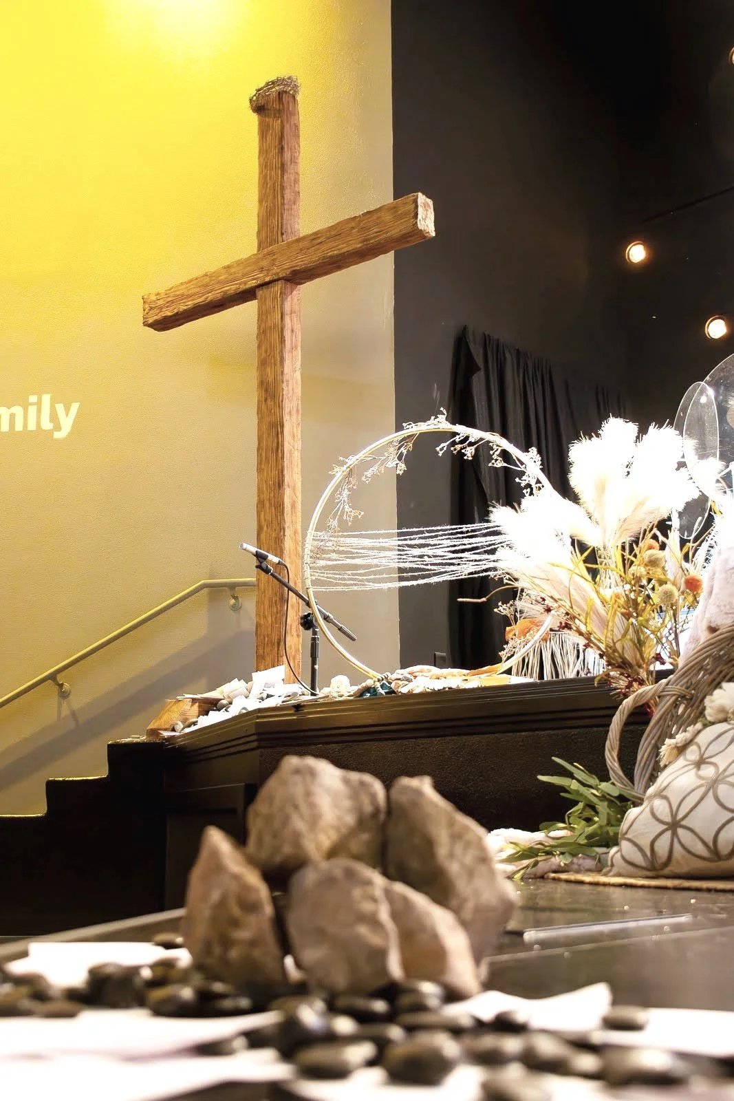 A wooden cross mounted on a wall with two different colored background walls, one yellow and one dark. In front of the cross, a table holds decorative dried flowers, a microphone, and shells or stones in the foreground.