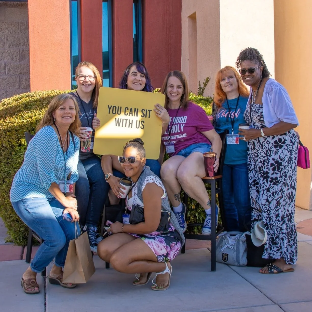 Group of seven women gathered outdoors, smiling, with some holding coffee cups. One woman in the middle holds a yellow sign that says, 'YOU CAN SIT WITH US.' They are standing or sitting in front of a building with red and yellow columns and greenery.
