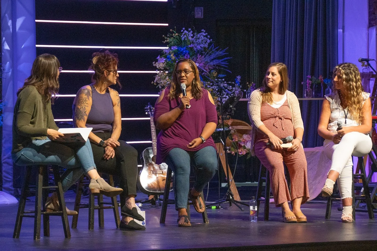 A panel of five women sitting on stools on a stage, engaged in a discussion. One woman is speaking into a microphone while others listen. The background features flowers and musical instruments.