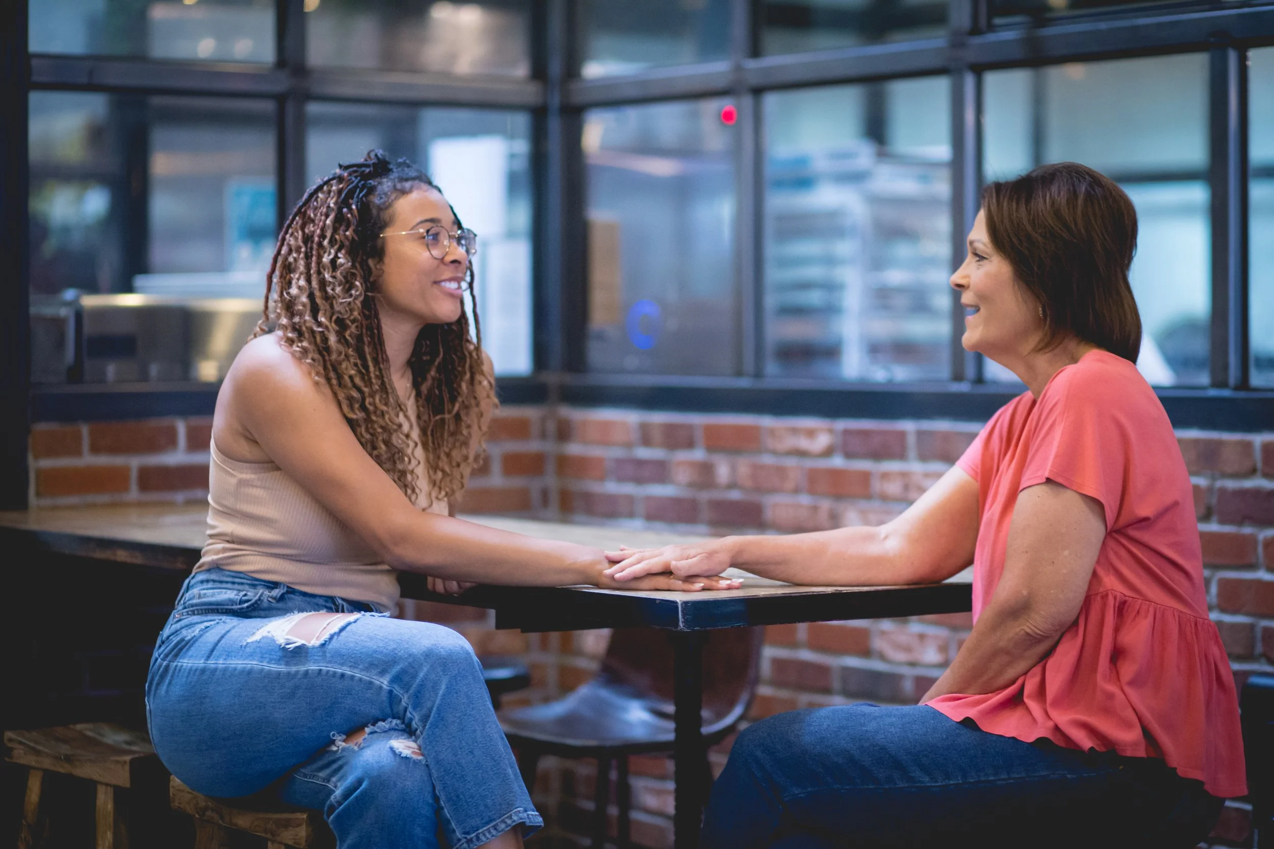Two women sitting at a table, holding hands, engaged in a heartfelt conversation in a cozy, modern cafe with brick walls and large windows.